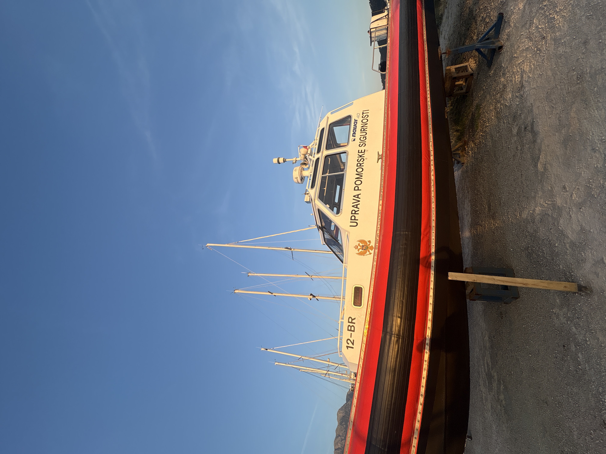 A color photograph depicts a boat with the words "UPRAVA POMORSKE SIGURNOSTI" (Maritime Safety Administration) written on the side, resting on supports. The hull is black with a red stripe. The number "12-BR" is painted on the side of the boat. In the background, there are masts from other boats. The sky is blue. The boat appears to be in a shipyard or dry dock. Based on the writing on the boat, this is likely in the city of Bar, Montenegro. The ground is covered in gray gravel or concrete.