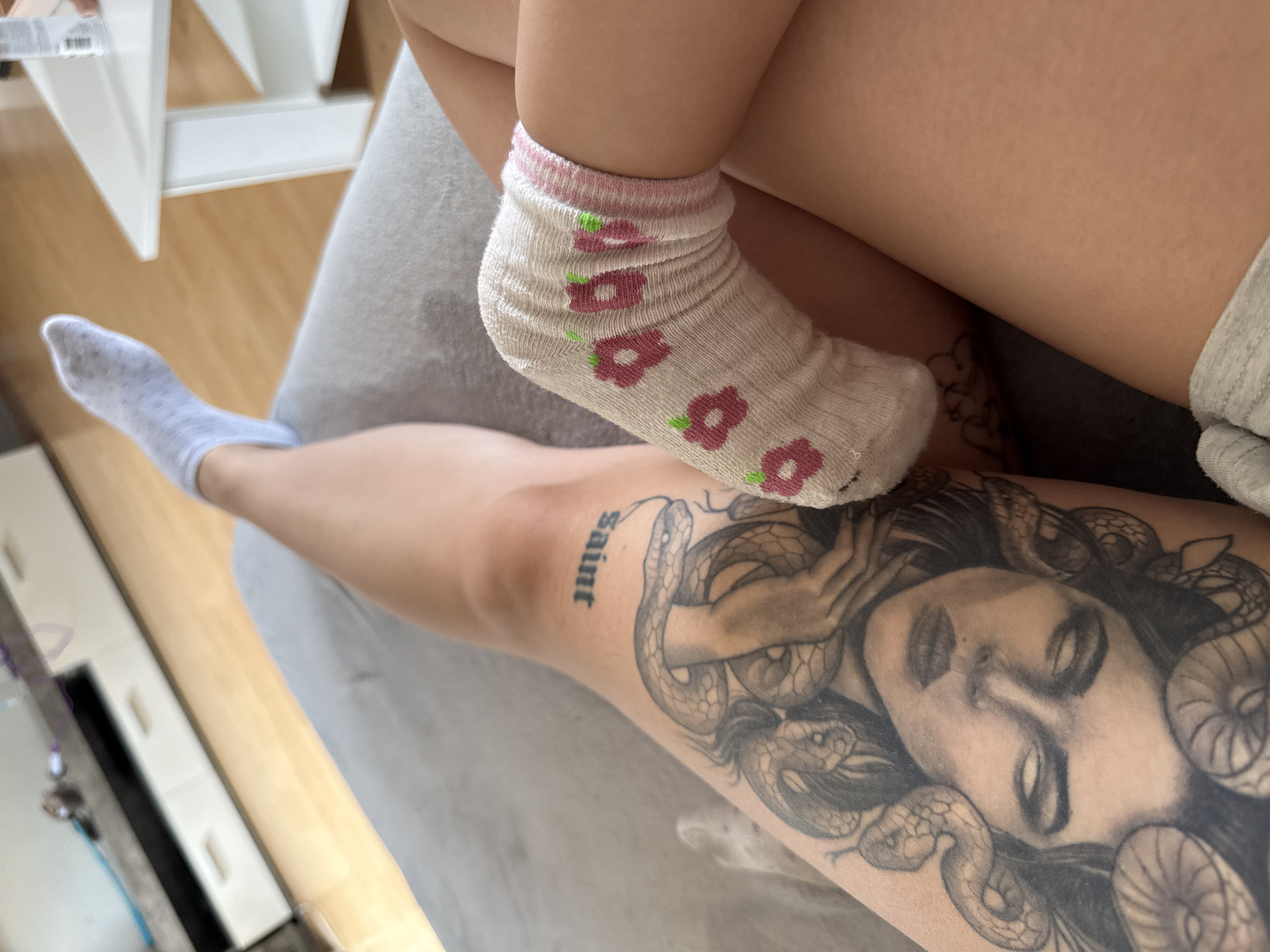 The image shows a close-up view of a person's legs and tattoos. The person is wearing a light blue sock on one foot and a white sock with pink flower patterns on the other. A large tattoo of Medusa with snakes for hair covers a portion of the thigh. The word "saint" is tattooed above the Medusa tattoo. The person is lying on a grey surface, possibly a sofa. In the background, a white coffee table and a wooden floor are visible. The environment appears to be indoors.