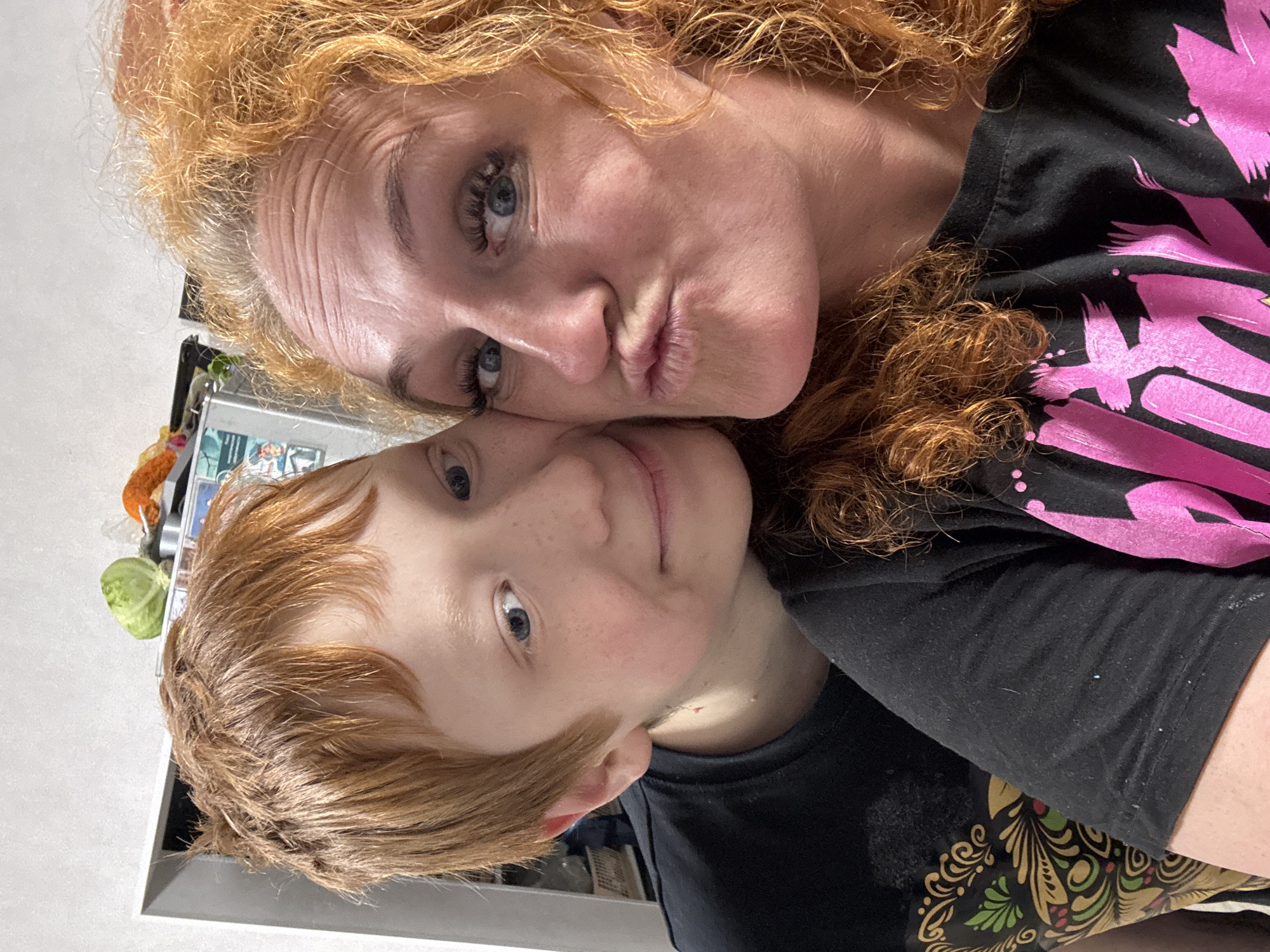 A selfie shows a woman and a child with red hair. The woman is making a kissing face and the child is smiling slightly. They are indoors. The environment appears to be a kitchen, based on the presence of a refrigerator and various objects on the counter. No information is available to verify the location.