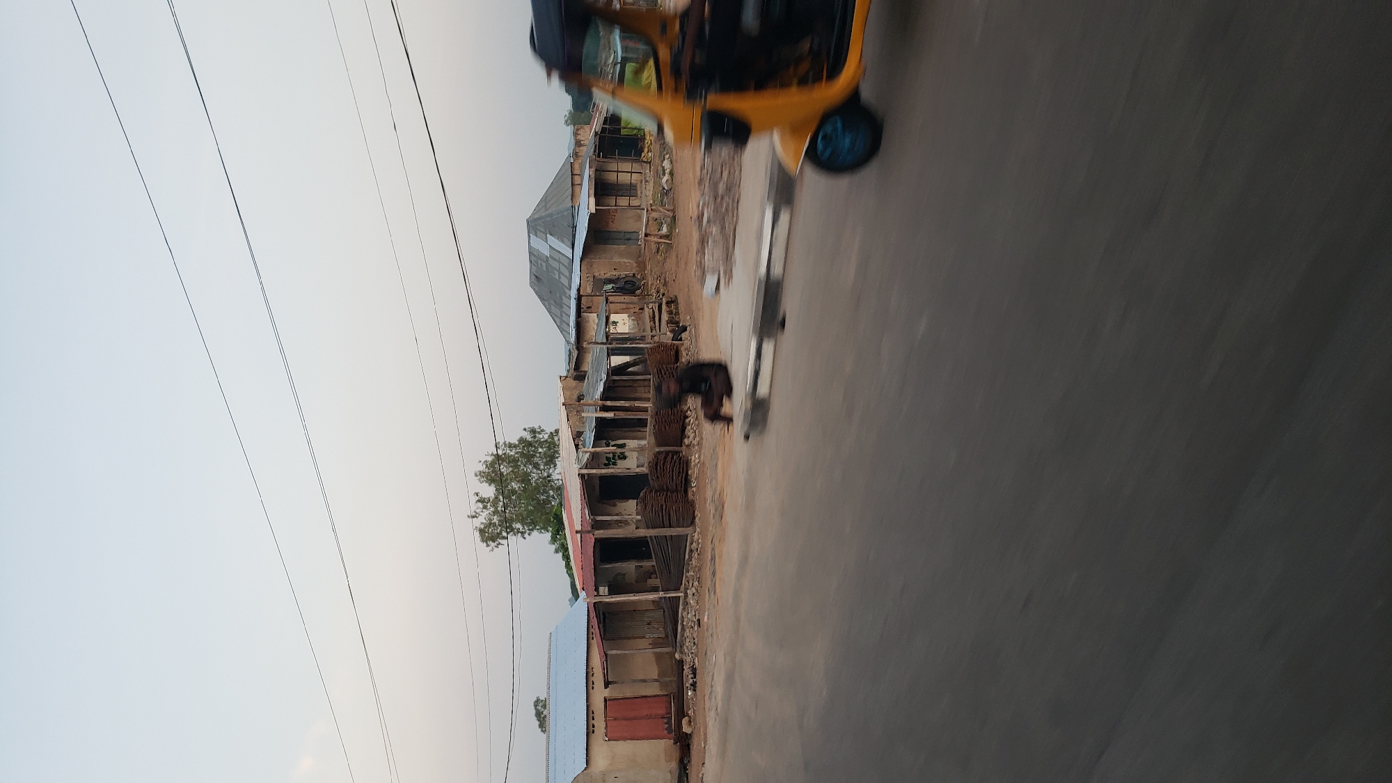 This is a photograph of a dusty street in Jalingo, Nigeria, likely in the late afternoon given the soft light and long shadows. The scene depicts a row of small, weathered buildings with corrugated metal roofs, some of which appear to be shops or residences. In front of the buildings, stacks of metal rods are piled up, possibly construction materials. A person is seated on the side of the road, facing away from the camera. In the foreground, a yellow auto-rickshaw (Keke Napep) is visible, blurred due to motion, suggesting the photograph was taken from a moving vehicle. The sky is a pale, hazy blue. Electrical wires stretch across the frame above the buildings. The ground is unpaved and appears dry and dusty, typical of the semi-arid climate in Jalingo.