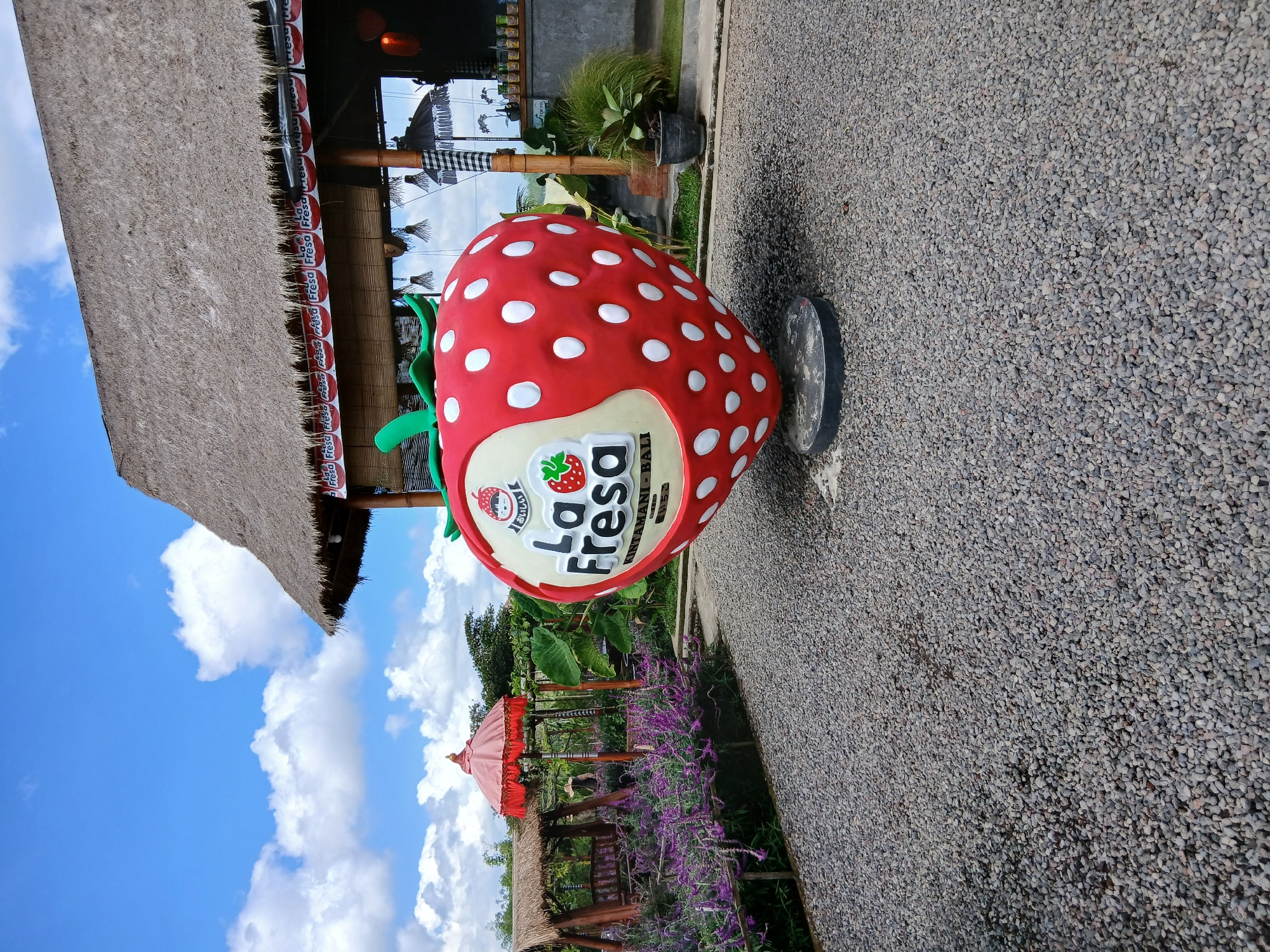The image shows a large, red, strawberry-shaped sculpture with white polka dots, prominently displayed on a gravel ground. The sculpture has a sign that reads "La Fresa" in large, black letters, with a smaller image of a strawberry and Japanese characters above it. Below "La Fresa" are the words "KINTAMANI BALI". In the background, a thatched-roof building with a rustic wooden structure is visible. This building appears to be a cafe or restaurant, with tables and chairs set up, and some decorative items like umbrellas. To the left of the strawberry sculpture, a garden area with vibrant purple flowers and a wooden swing set can be seen. The sky is blue with scattered white clouds. The overall scene depicts an outdoor recreational area, likely a tourist attraction, in Kintamani, Indonesia, characterized by its agricultural theme, with the strawberry being a central motif.