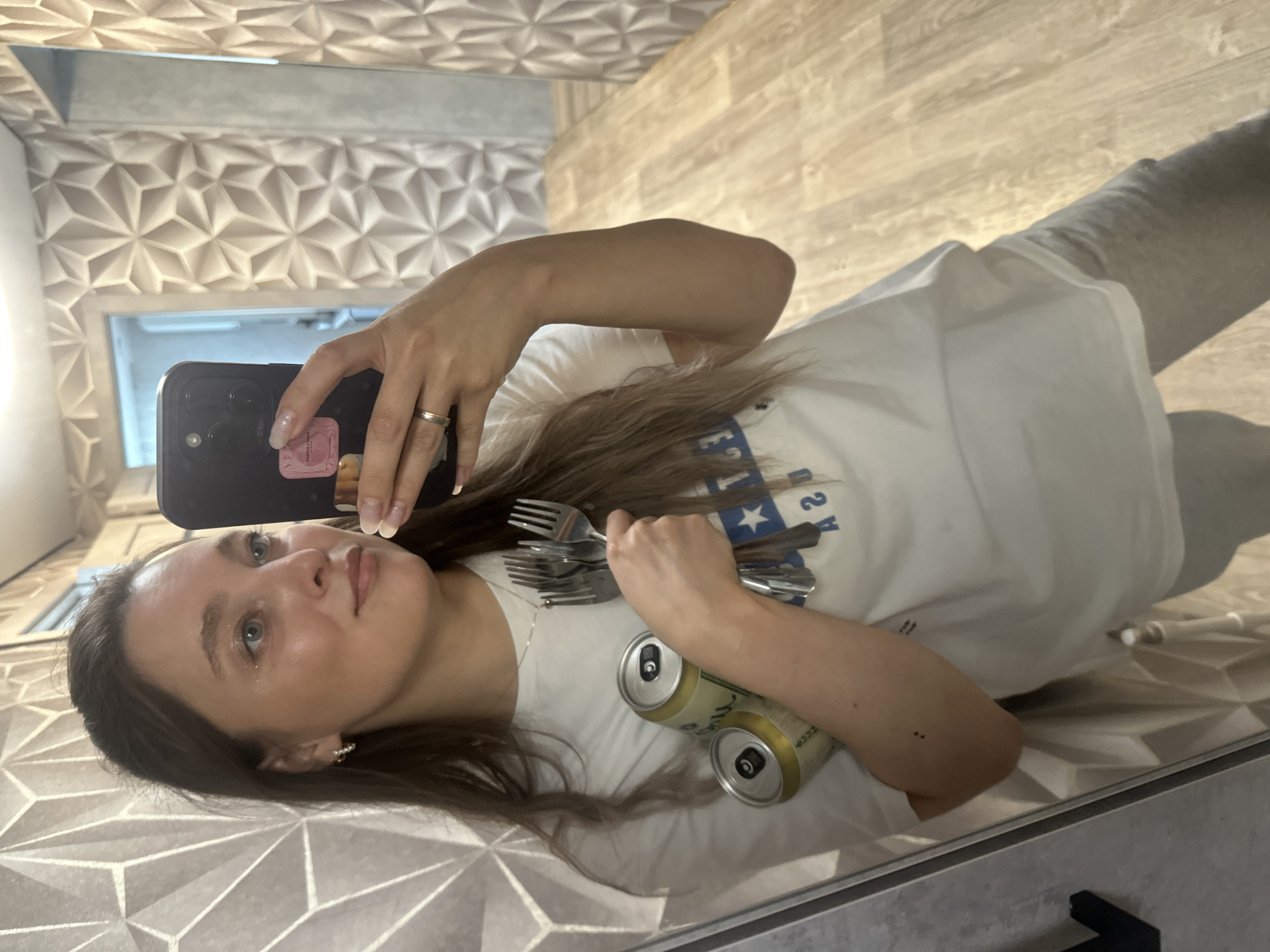 A young woman with light brown hair, wearing a white t-shirt and grey sweatpants, is taking a mirror selfie. She is holding two cans of what appear to be beer and a bunch of forks in her left arm. She is also wearing a silver ring on her left ring finger and small hoop earrings. Her face is turned towards the camera, and she is smirking slightly. In the background, there is a wall with a geometric textured pattern and a wooden floor. The scene appears to be indoors, possibly in a bathroom or hallway. There is no indication of the city of Вознесенка, Russia.