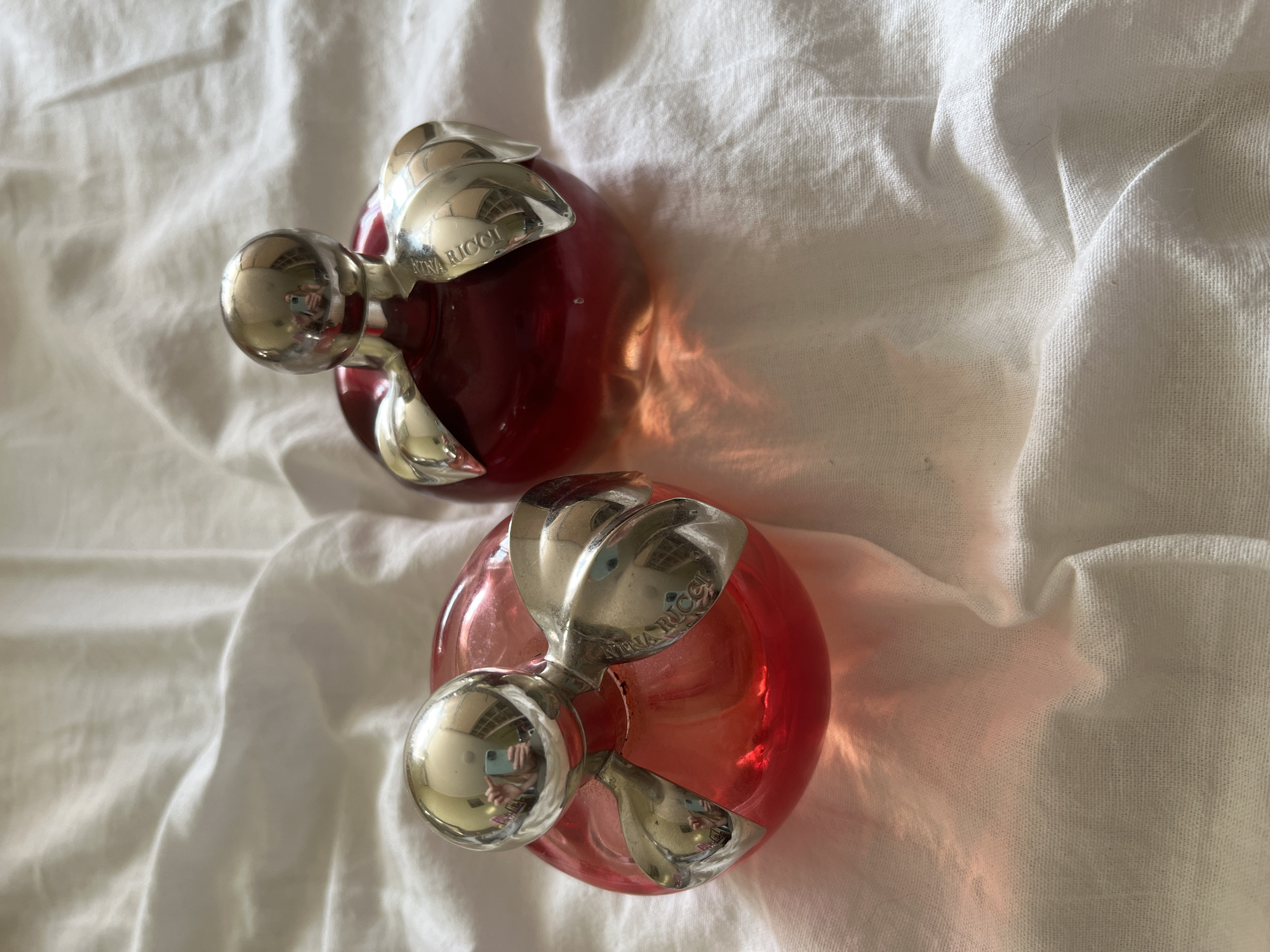 The image shows two red, apple-shaped perfume bottles with silver caps and leaf-like accents, placed on a white, wrinkled fabric surface. The bottle on the right has "NINA RICCI" engraved on one of the silver leaves. The lighting suggests an indoor setting, possibly a bedroom or vanity. There is no indication of the city of Komsomolskoye, Ukraine, or any people or actions taking place.