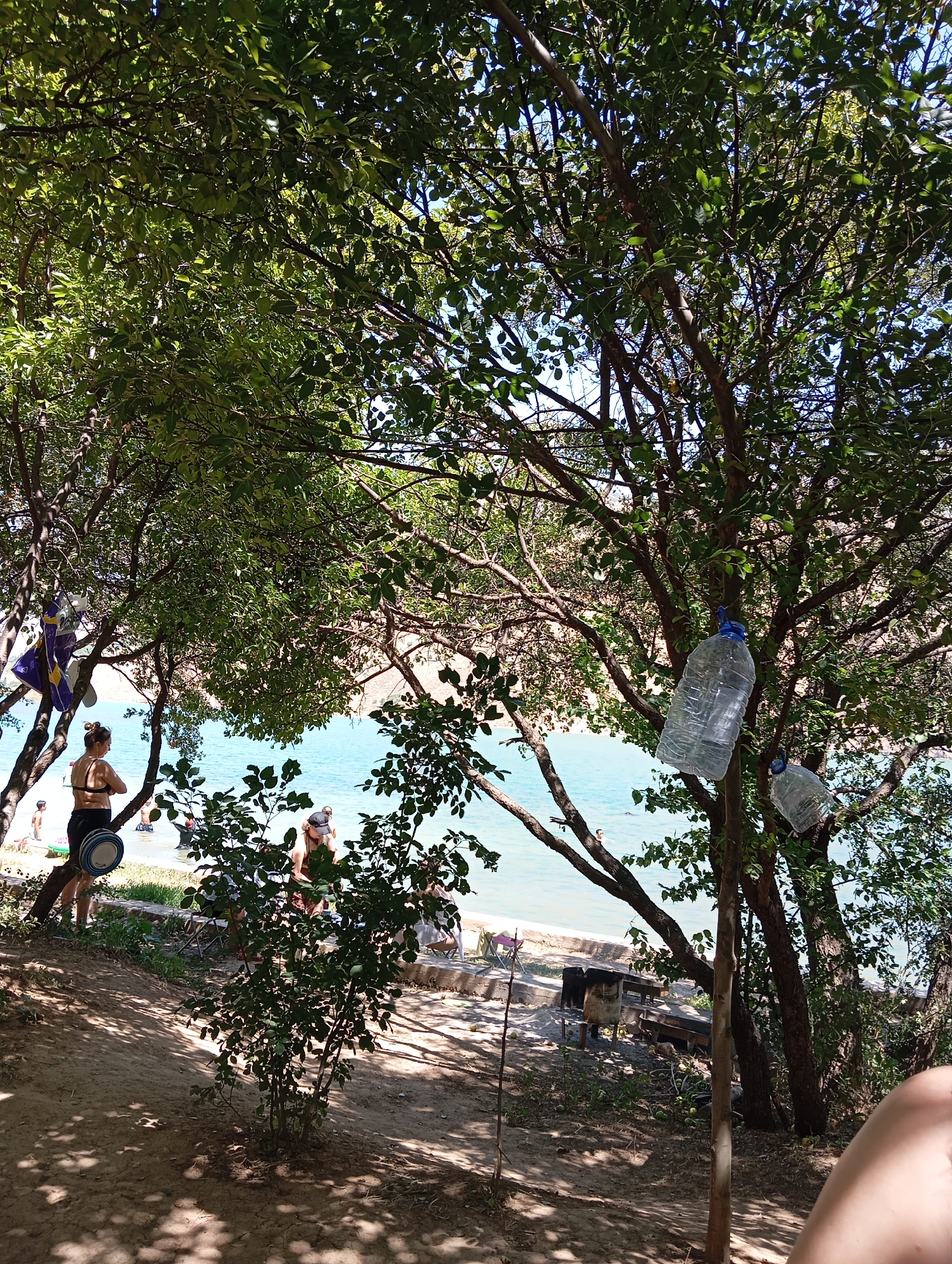 The image depicts a scene on the shore of a body of water, possibly a lake or river, in the city of Yangikurgan, Uzbekistan. A group of people are present, some wading in the water and others relaxing on the sandy shore under the shade of trees. In the foreground, a woman in a black bikini is standing by the water's edge, facing away from the camera. She has a hat in her hand. To her left, a child is partially visible. Further down the shore, several other individuals are seen, some in swimsuits. Large plastic bottles are hanging from tree branches, one prominently in the mid-right of the frame. The area appears to be a recreational spot, with a sandy beach, vegetation, and trees providing shade. The water is a bright blue, and the sky is clear. There are some items of camping or picnic equipment visible, including a grill or barbecue setup. The overall impression is a relaxed, outdoor gathering by the water on a sunny day.