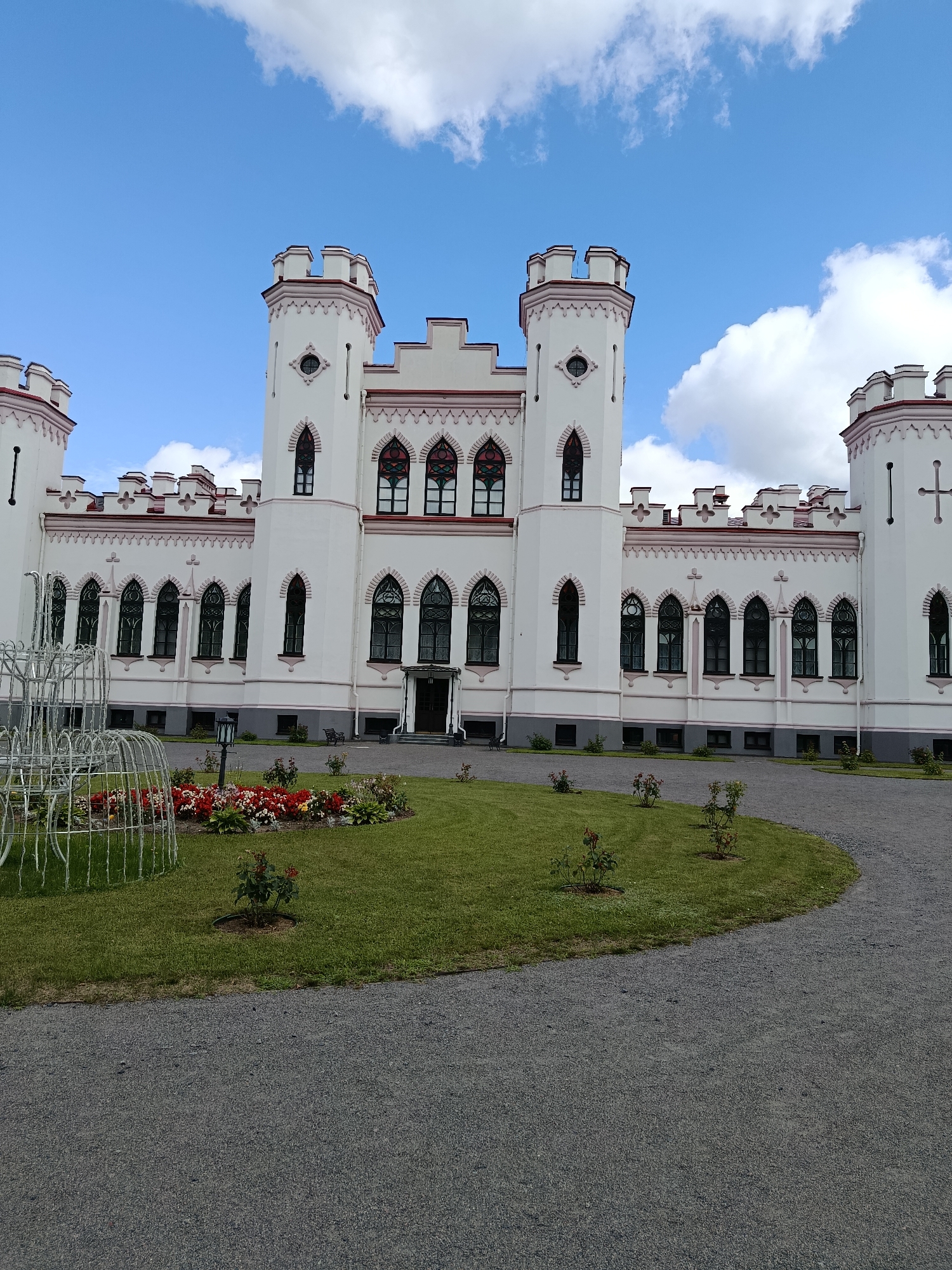 The image shows a white, Gothic-style castle with red trim, featuring crenellated towers and arched windows. In front of the castle is a well-maintained lawn with a flowerbed of red and white flowers and several small rose bushes. A gravel driveway curves around the lawn. A white, open-cage-like structure is visible on the left side of the frame. The sky is blue with scattered white clouds. The castle is identified as Rumyantsev-Paskevich Palace in the village of Kvasyavichy, Квасевіцкі сельскі Савет, Belarus.