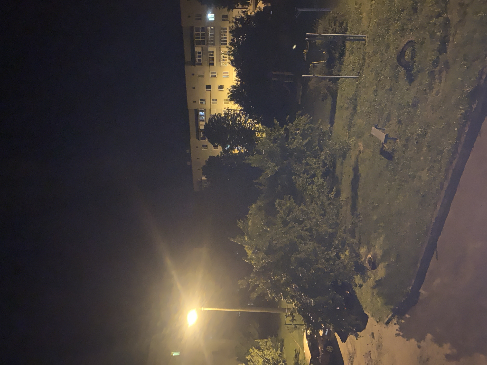 The image depicts a nighttime scene in a residential area of Sumy, Ukraine. A tall, illuminated street lamp dominates the left side of the frame, casting a warm glow on the surrounding trees and a portion of a road. In the background, a multi-story apartment building with lit windows is visible. In the foreground, a patch of grass features a simple wooden bench and what appears to be a discarded tire. The overall environment suggests a typical urban setting at night, with elements of street lighting, residential architecture, and a small public green space.