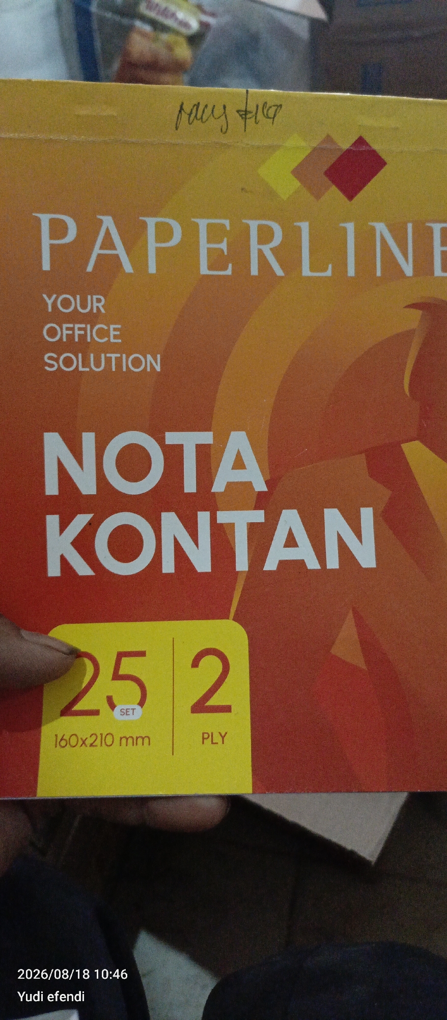 The image displays a close-up of a product box. The top portion of the box has handwritten text that appears to say "racy fig". Below this, the prominent branding "PAPERLINE" is visible, followed by "YOUR OFFICE SOLUTION" in smaller white text. Further down, large white text reads "NOTA KONTAN". At the bottom left of the box, a yellow rectangle contains bold red numbers "25" and "2", with the text "SET", "160x210 mm", and "PLY" underneath. The background of the box features a gradient of orange and red, with abstract circular patterns. In the bottom right corner of the image, a timestamp "2026/08/18 10:46" and the name "Yudi efendi" are present, suggesting this image was taken on that date and time, attributed to Yudi efendi. The image is not of a scene in Rawajitu Selatan, Indonesia.