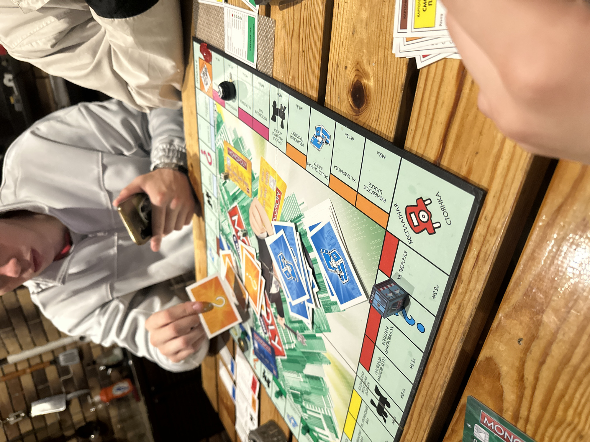 The image shows a group of people playing a board game, likely Monopoly, around a wooden table. The game board is spread out, with various property cards, dice, and game pieces visible. One person is holding a card with a question mark on it, appearing to be interacting with the game. Another person is holding a mobile phone. The environment appears to be indoors, possibly a home or a casual gathering space, with a brick wall and kitchen elements visible in the background. The game board features property names written in Cyrillic script, indicating a Russian version of the game. Specifically, properties like "ул. Тверская" (Tverskaya Street) and "Площадь Маяковского" (Mayakovsky Square) are visible, suggesting a setting within Russia, potentially Moscow. The game is in progress, with visible game tokens and dice indicating movement and strategic play. The overall atmosphere suggests a social gathering centered around a board game.