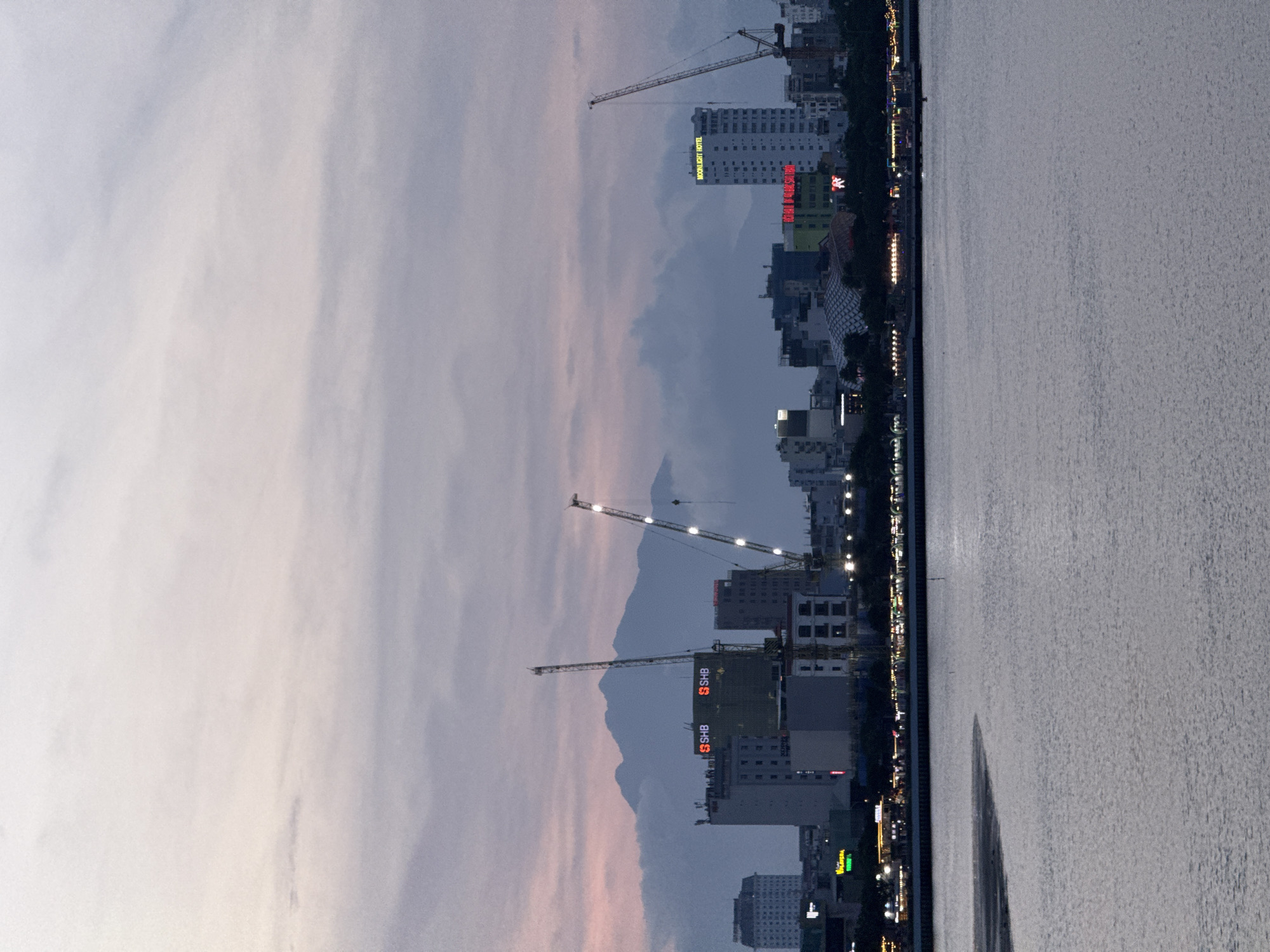 The image captures a panoramic view of the city of Da Nang, Vietnam, from across the Han River at dusk. In the foreground, the Han River's surface reflects the muted colors of the twilight sky. The middle ground features a cityscape dotted with various buildings, including several high-rise structures. Prominent among these are buildings with illuminated signage. Two construction cranes are visible, one on the left and another on the right, indicating ongoing development. In the background, a mountain range is silhouetted against the sky, partially obscured by clouds. The sky itself is a canvas of soft, pastel hues of pink, purple, and gray, characteristic of sunset or sunrise. The lighting suggests it is either early morning or late evening.
