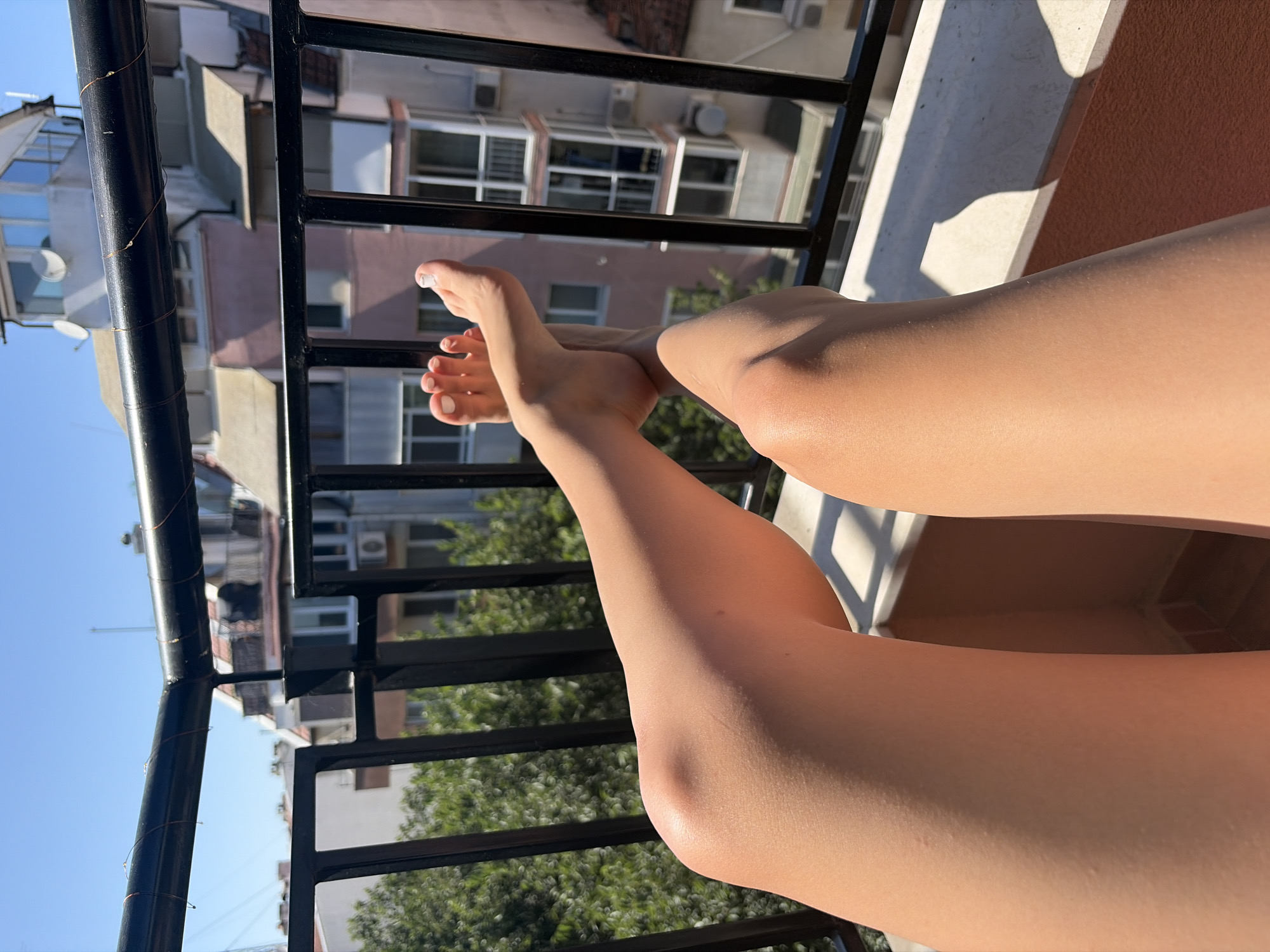 The image shows a person's bare legs and feet resting on a balcony railing in Burgas, Bulgaria. The person appears to be relaxing in the sun, with the lower half of their body in the foreground. Their legs are tanned and smooth, and their feet are neatly pedicured with white nail polish. In the background, several multi-story buildings are visible, with balconies, windows, and air conditioning units. Trees and a clear blue sky complete the urban landscape. The overall impression is one of a warm, sunny day in a city.