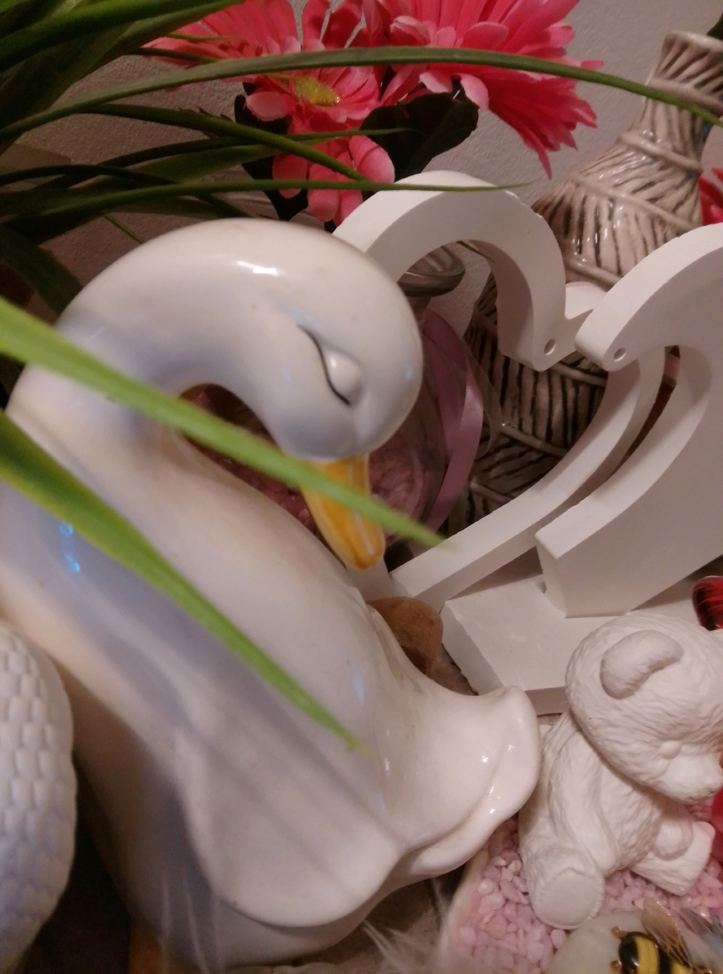 The image depicts a close-up of a decorative arrangement. In the foreground, a white ceramic swan sculpture is positioned on the left. Its head is slightly bowed, and a yellow beak is visible. Green leaves partially obscure the swan. To the right of the swan, a small, unpainted white ceramic bear sculpture is seated on a bed of small pink stones. A decorative object, possibly a vase with a striped pattern, is visible in the background, along with pink faux flowers. A white heart-shaped decorative element is also present. The lighting suggests an indoor setting, likely a home decoration. This scene is not located in the city of Wedel, Germany.