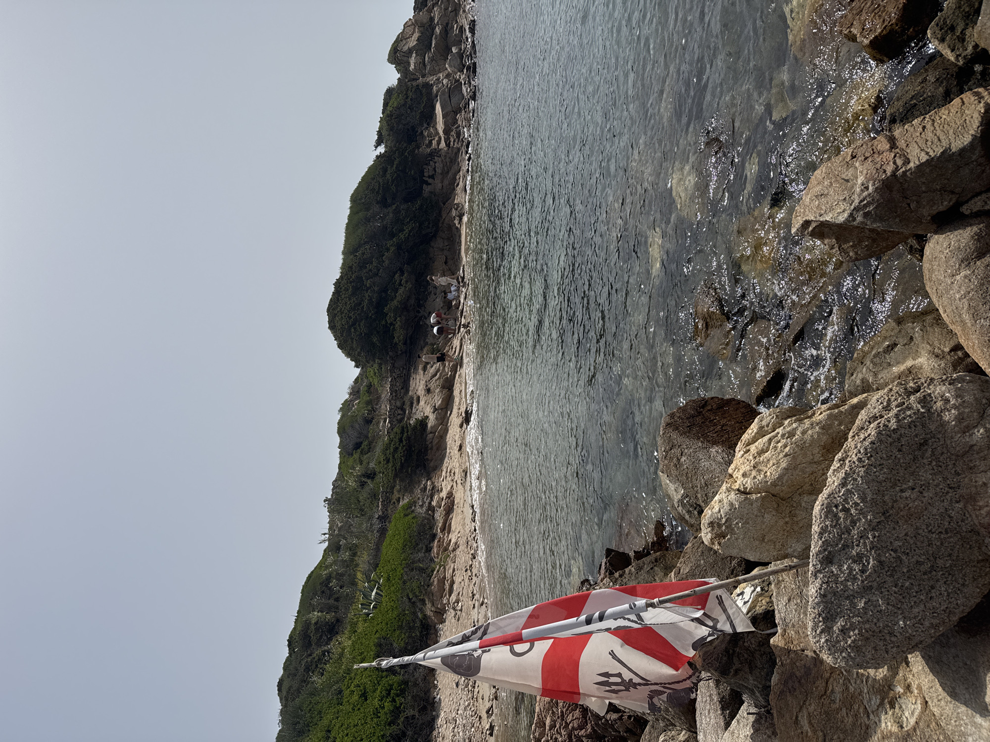The image depicts a coastal scene in La Maddalena, Italy. The foreground is dominated by a flag with red and white stripes and a black trident symbol. The flag is attached to a pole and appears to be resting on large rocks. Behind the flag, a narrow beach meets the clear, shallow water of the sea. The coastline is lined with lush green vegetation and rocky outcrops. In the mid-ground, a few people are visible on the beach, appearing to be engaged in leisurely activity. The sky is overcast, suggesting a cloudy or hazy day. The overall impression is one of a quiet, natural setting, possibly a secluded cove or bay. The lighting is diffused, indicating it's likely daytime. There is no visible text in the image.