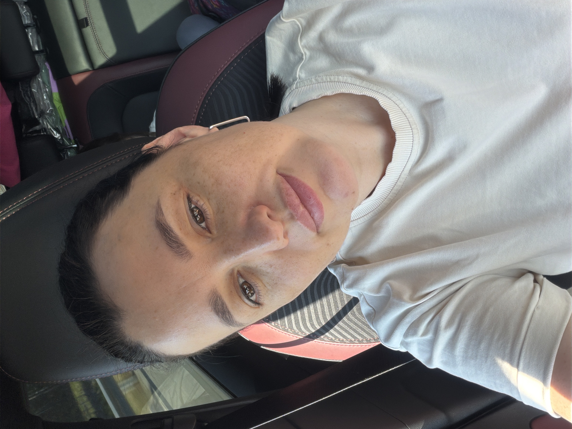 A woman with brown hair and brown eyes, wearing a light-colored t-shirt and square earrings, is taking a selfie in a car. The car's interior, featuring dark leather seats with red stitching, is visible. Sunlight casts shadows across the scene. The woman has freckles on her face. The location is identified as Uvarovo, Russia.