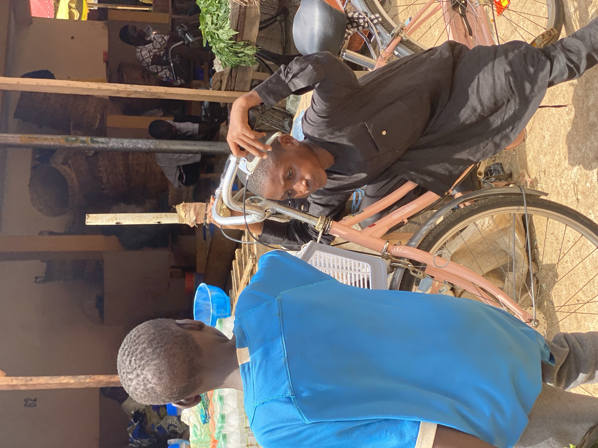 The image shows an outdoor scene in Kafanchan B, Nigeria. A person wearing a black shirt is sitting on a pink bicycle, holding a metal object to their head. A second person, wearing a bright blue shirt, stands in front of the bicycle. The background includes a building with wooden supports, stacked items, and other people further away. Various objects like vegetables and containers are visible. The lighting suggests it is daytime.