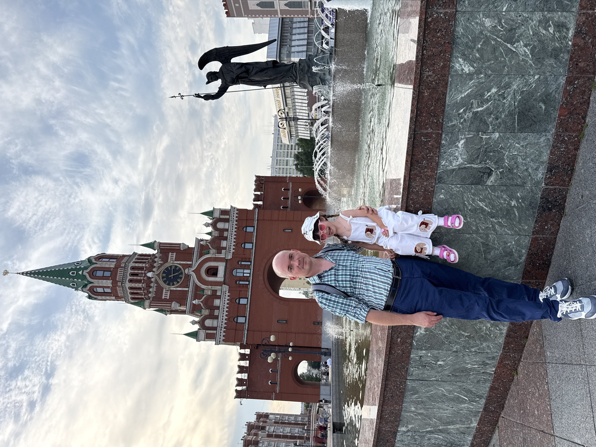 In Yoshkar-Ola, Russia, a bald Caucasian man and a young Caucasian girl are posing for a photo in front of a fountain and a tower. The man, wearing a plaid shirt and blue pants, stands to the left, while the girl, dressed in white overalls and a hat, sits to his right. In the background, a tall, red brick tower with a green spire and a clock face stands prominently. To the right of the tower, a large fountain with water jets is visible, and beyond that, a dark statue of an angel with outstretched wings holding a spear. The scene is set against a cloudy sky.
