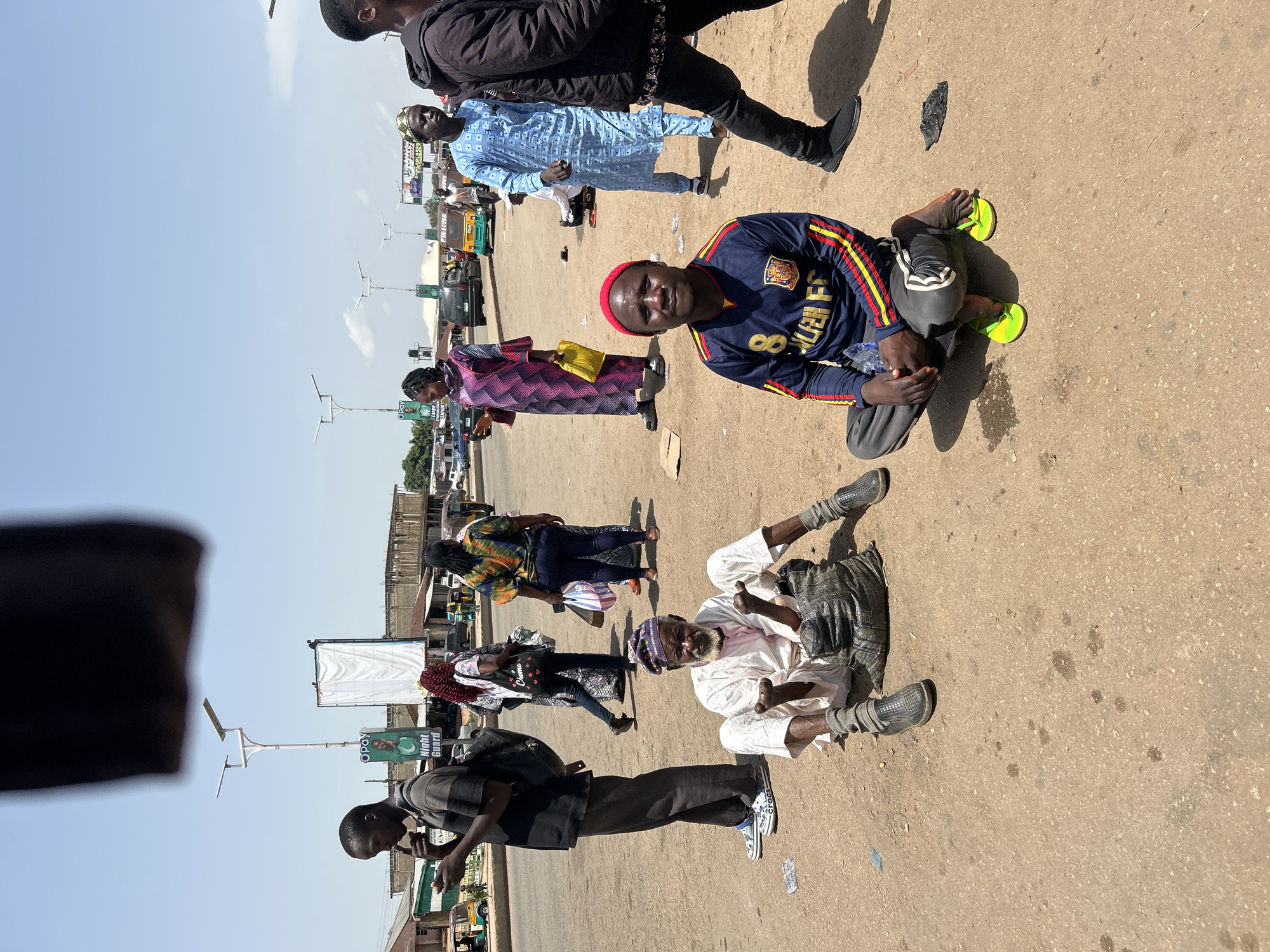 The image depicts a street scene in Jos, Nigeria. In the foreground, a man sits on the ground wearing a blue and red long-sleeved shirt with the number 8 and the letters "FC" visible, along with gray pants and bright green flip-flops. He is looking towards the camera. To his left, an older man sits on the ground, wearing traditional white clothing and a patterned cap, with his hands held out. He is positioned next to a dark-colored sack. Behind them, several other individuals are walking or standing. A woman in a colorful patterned dress and a headscarf walks past, carrying a yellow bag. Another woman in a patterned top and dark pants also walks by. A man in a blue kaftan with a patterned design walks further back. Further in the background, there are vehicles including a tricycle and cars on a dusty road, with buildings and streetlights lining the area. The sky is bright blue with a few white clouds.