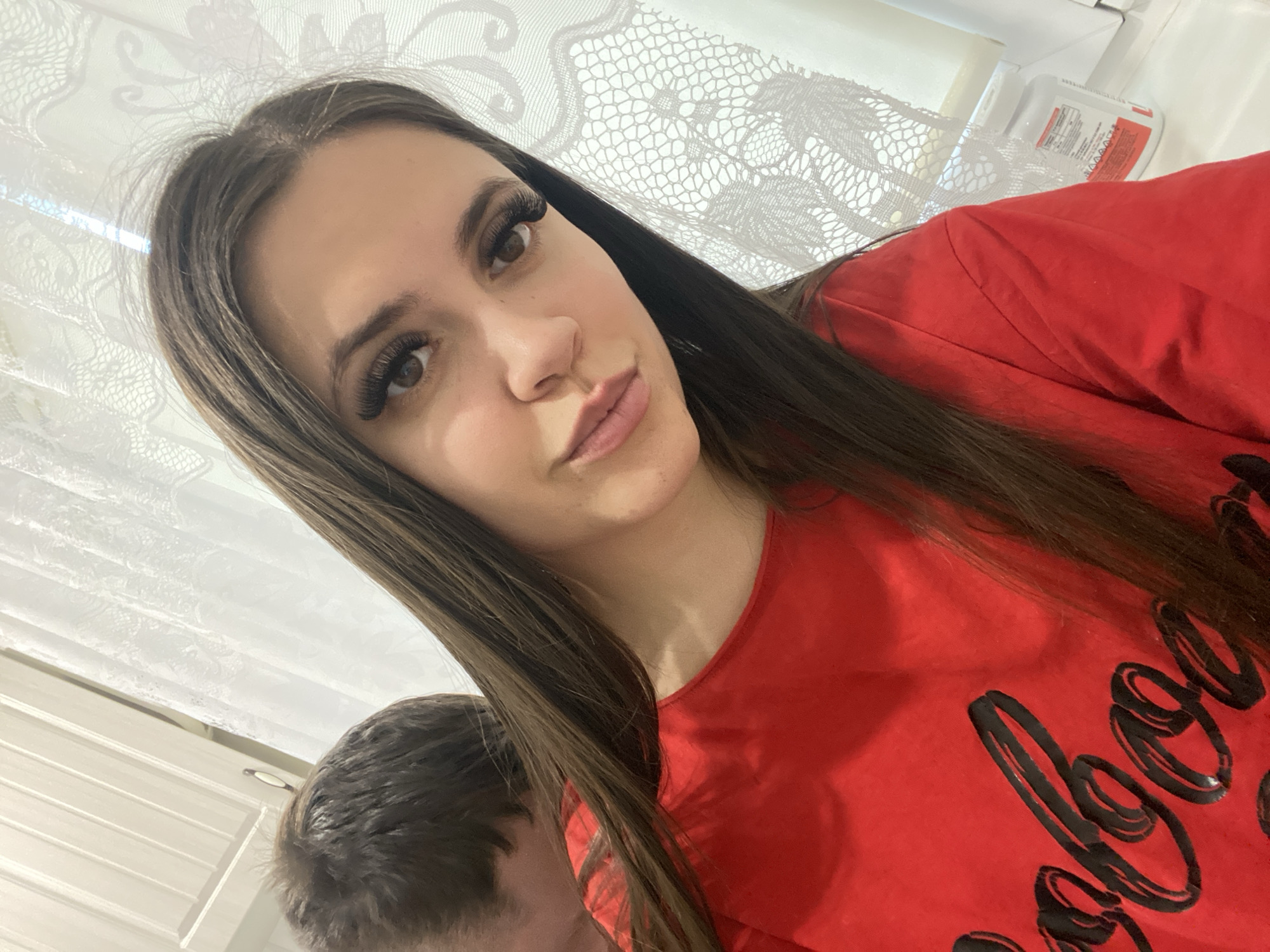 A young woman with long, straight dark brown hair and heavy eyelashes is posing for a selfie. She is wearing a red t-shirt with black writing. A portion of another person's head is visible in the lower left corner. Behind the woman, there is a white lace curtain. In the background, there is a white cabinet and a white bottle. The location appears to be indoors. Given the user prompt, it is theoretically located in the city of Chaikovsky, Russia.