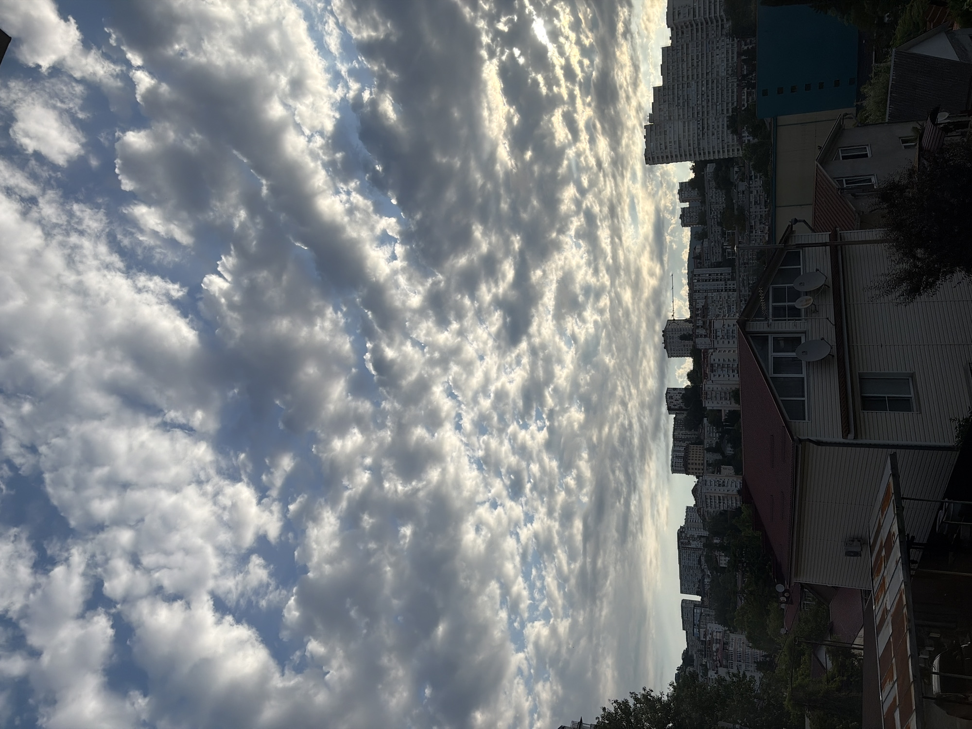 This is a landscape photograph taken from a high vantage point, looking out over a city with a cloudy sky. The foreground features the roof of a house with a red roof and some satellite dishes mounted on its exterior. Behind it, more houses and buildings of varying sizes are visible, with trees scattered throughout. The middle ground is dominated by a dense cluster of apartment buildings, many of which are tall and modern, built on a hillside. A large, distinctive curved apartment building is prominently featured on the right side. The background consists of a dramatic sky filled with cumulus clouds, some of which are lit by the sun breaking through, creating a sense of depth and texture. The overall scene suggests an urban environment, likely in a coastal or hilly region. Based on the distinctive architecture of the buildings, particularly the curved apartment complex, and the hilly terrain, this image is likely taken in the city of Sochi, Russia. There are no people visible in the frame, and no actions or interactions are depicted. The environment is characterized by residential and multi-story buildings, lush greenery from trees, and a dynamic cloudscape.