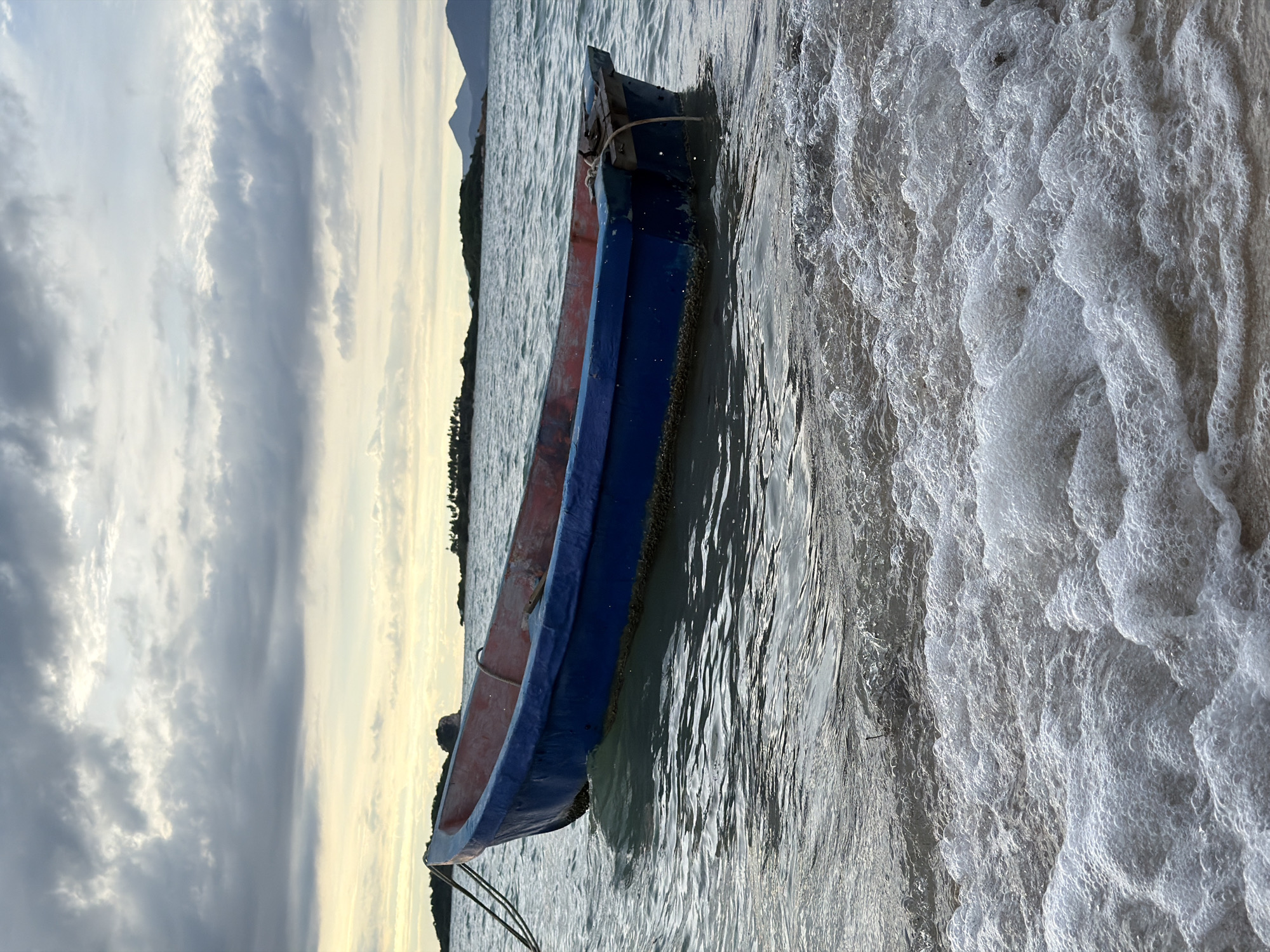 A small, weathered blue and red boat rests partially in the water near a shore covered in frothy white seafoam. The boat appears to be made of wood and has a rope tethered to its front. The water is choppy, with sunlight glinting off the surface and creating ripples. In the background, a distant shoreline with green vegetation and faint mountains can be seen under a sky filled with dramatic, grey clouds tinged with yellow light, suggesting either sunrise or sunset. The overall scene is tranquil, with the gentle motion of the waves and the muted colors of the sky creating a peaceful atmosphere. The location appears to be a beach in Baan Plai Laem, Thailand, indicated by the tropical vegetation and mountainous terrain visible in the distance. There is no visible text in the image.