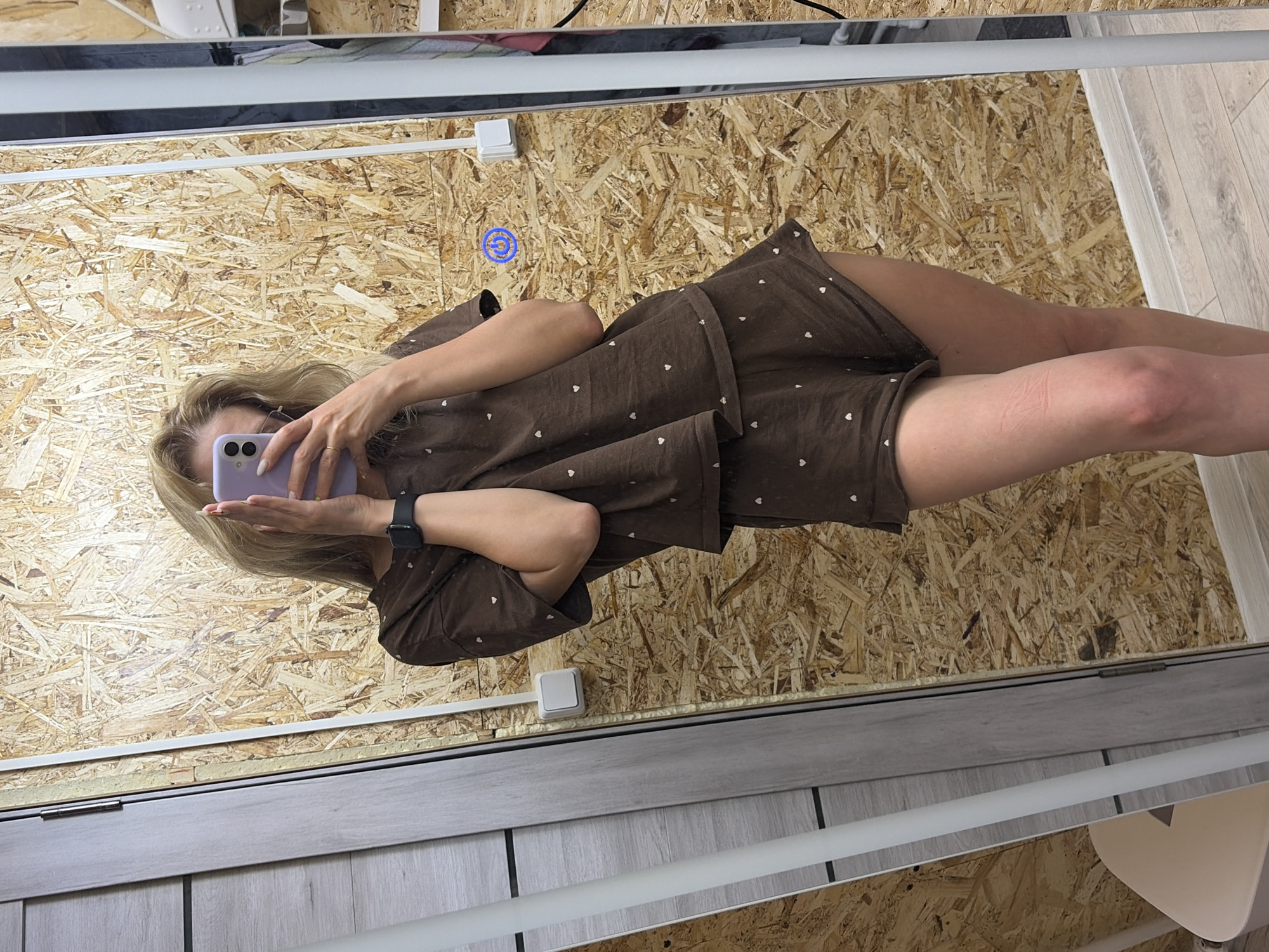 A woman is taking a mirror selfie. She has blonde hair and is wearing a brown, patterned pajama set and a black smartwatch. She is holding a lavender-colored cell phone. The background is a wall made of wood particle board. There are light switches on the wall, and the reflection of a white ceiling fixture is visible in the mirror. The floor is light wood-tone planks. No location is confirmed or confirmed from this image.