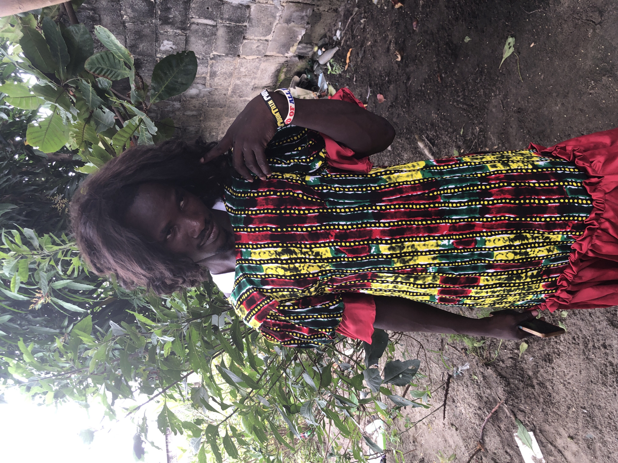 A person with dark, wavy hair is standing outdoors in what appears to be a somewhat unkempt environment. They are wearing a brightly colored dress with a pattern of vertical stripes in red, yellow, green, and black, accented with small yellow dots. The dress has puffy sleeves and a ruffled hemline in red. The person is holding a mobile phone in their left hand and has their right hand near their head, touching their hair. They are also wearing two wristbands on their right arm, one of which appears to have text on it. In the background, there are lush green trees and a brick wall. The ground is covered in dirt and scattered debris. The lighting suggests it is daytime. The scene is located in Ado, Nigeria.