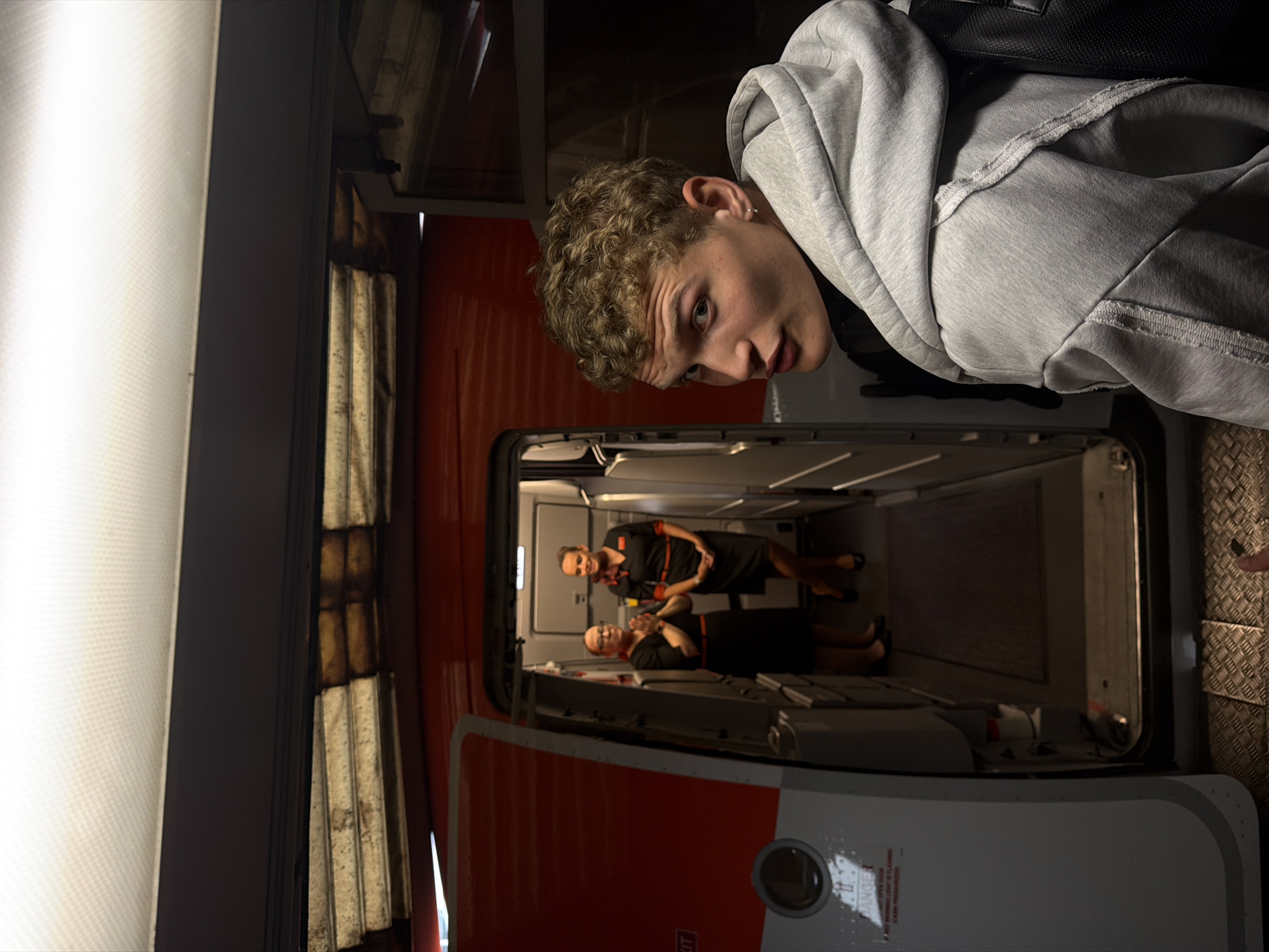 A young man with curly blonde hair is seen from the shoulders up in the foreground, looking over his right shoulder towards the camera. He is wearing a grey hooded sweatshirt. Behind him, two women in flight attendant uniforms are visible through an open airplane door. The woman on the left is wearing glasses and has her hands clasped in front of her. The woman on the right is smiling. The interior of the airplane, with red accents, is visible. The scene appears to be at an airport in Crawley, United Kingdom.
