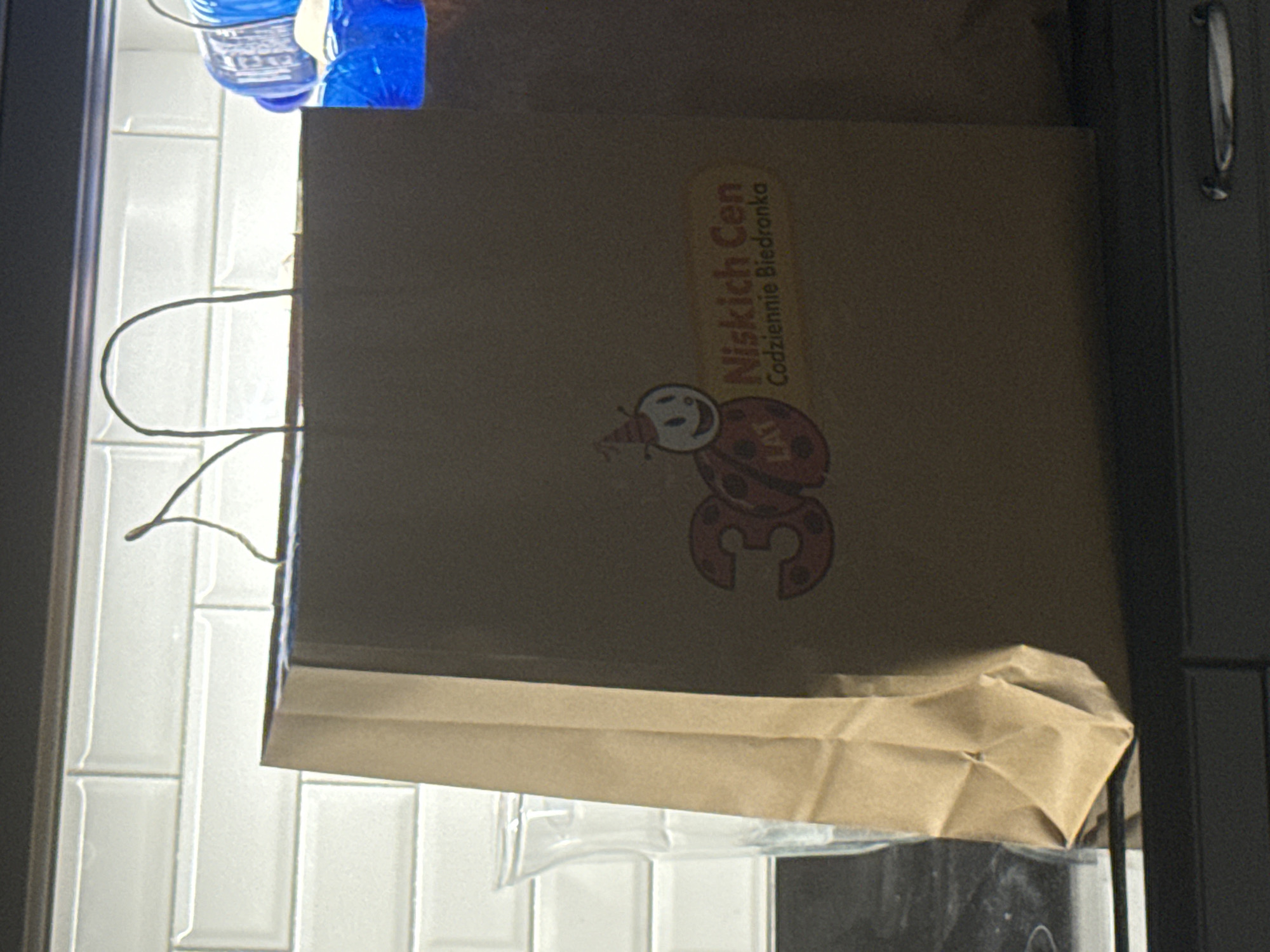 The image shows a brown paper shopping bag with the logo "Niskich Cen Codziennie Biedronka" printed on it, along with a cartoon ladybug character wearing a party hat and the number "30" with polka dots. The bag is sitting on a dark countertop, with a clear glass vase to its left. Behind the bag is a white subway tile backsplash. To the right of the bag, another shopping bag is partially visible, and a blue plastic bottle with a white cap is leaning against it. The scene appears to be in a kitchen or dining area.