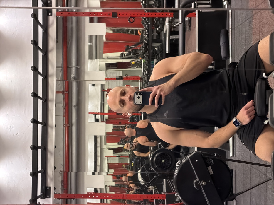 A bald man in a black tank top and shorts sits on a rowing machine, holding a phone up to take a selfie. He wears a smartwatch. Behind him is a mirrored wall with multiple reflections of him. The gym has red and black equipment. He has an earbud in one ear.