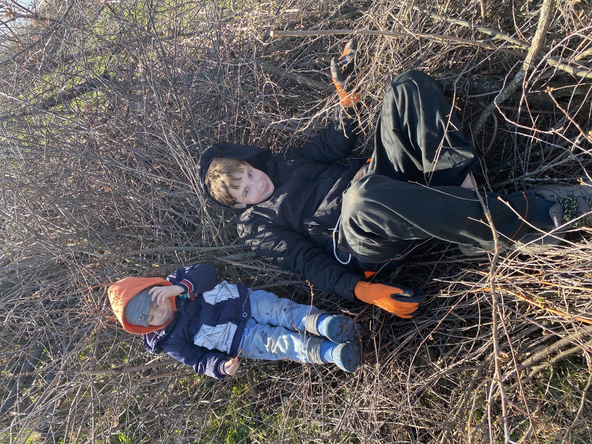 Two children lie amongst a dense thicket of dry, thorny branches, possibly in a natural area within the city of Ольховка, Russia. The older child is wearing a dark-colored jacket, dark pants, orange gloves, and hiking shoes. They are lying on their back and looking toward the camera, holding a stick. The younger child wears an orange hooded sweatshirt over a gray hat, a dark blue and white jacket, light blue pants, and blue boots. They appear to be resting or playing among the branches. The environment consists primarily of intertwined bare branches and twigs. Some green grass and vegetation are visible in the lower left corner.