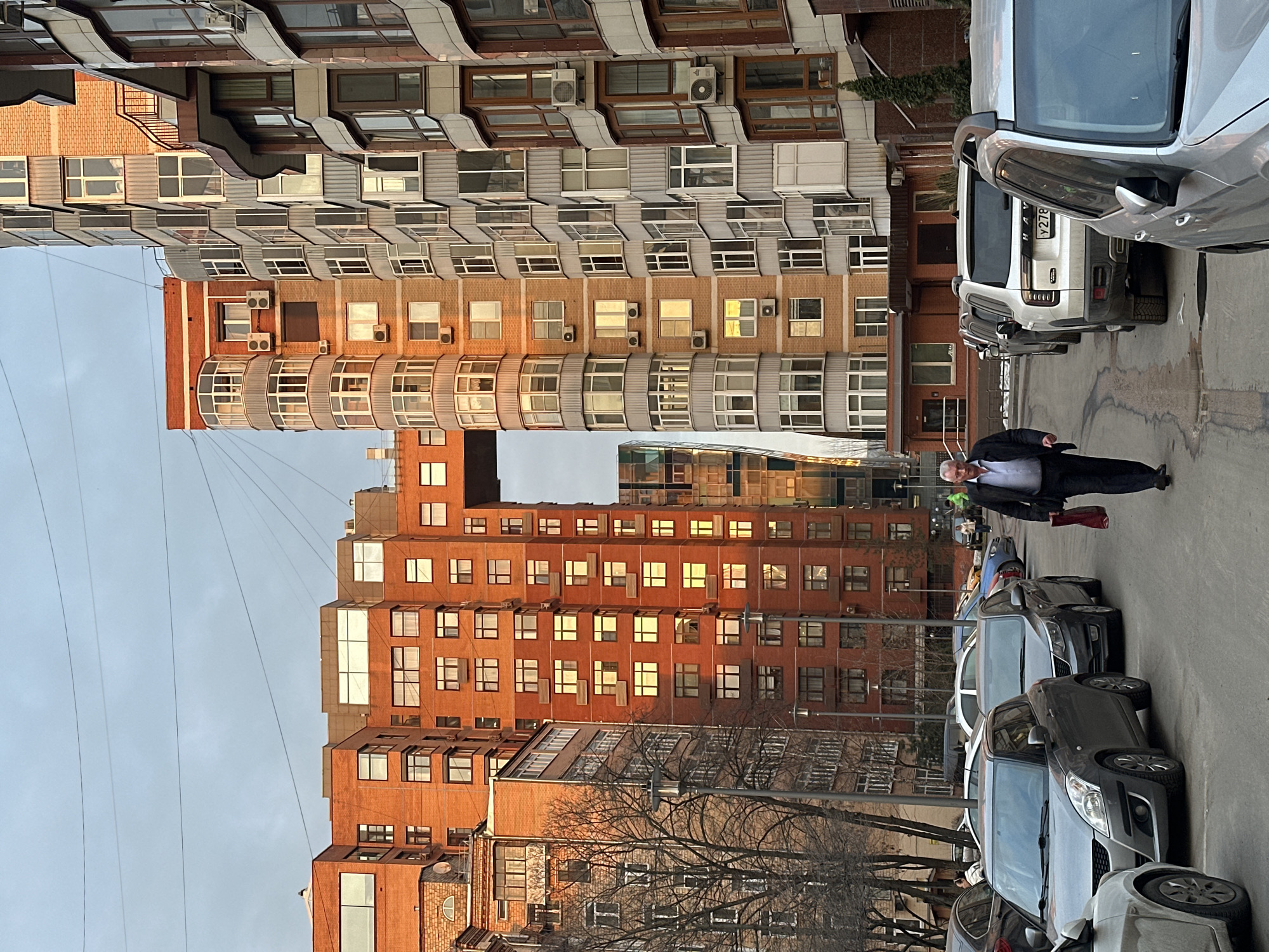 An urban street scene depicts the residential area surrounding the House on Mosfilmovskaya complex at Mosfilmovskaya Street 8, Ramenki District, Moscow, Russia. The image is captured during late afternoon or early evening, indicated by golden sunlight illuminating building facades against a hazy, overcast sky.
The dominant structures are multi-story residential buildings. On the left, a tall red-brick building, part of the House on Mosfilmovskaya complex, features numerous rectangular windows and a distinctive elevated walkway connecting two sections. Its western facade is bathed in warm, orange sunlight. To the right, another modern, tall residential building presents a complex facade of grey panels, brickwork, and numerous curved bay windows and balconies, with multiple outdoor air conditioning units visible.
The ground level consists of a paved asphalt surface, showing wet patches or shallow puddles. Vehicles are parked in rows along both sides of the thoroughfare. Notable cars include a grey Suzuki Swift hatchback, a dark sedan, and a white Haval SUV displaying a Russian license plate (Y278). Bare deciduous trees are scattered along the left side of the street.
A single older Caucasian male, wearing a dark jacket, light blue collared shirt, and dark trousers, walks towards the viewer, slightly right of the central axis. He carries a red bag in his left hand. Additional individuals are minimally visible in the background near parked vehicles.