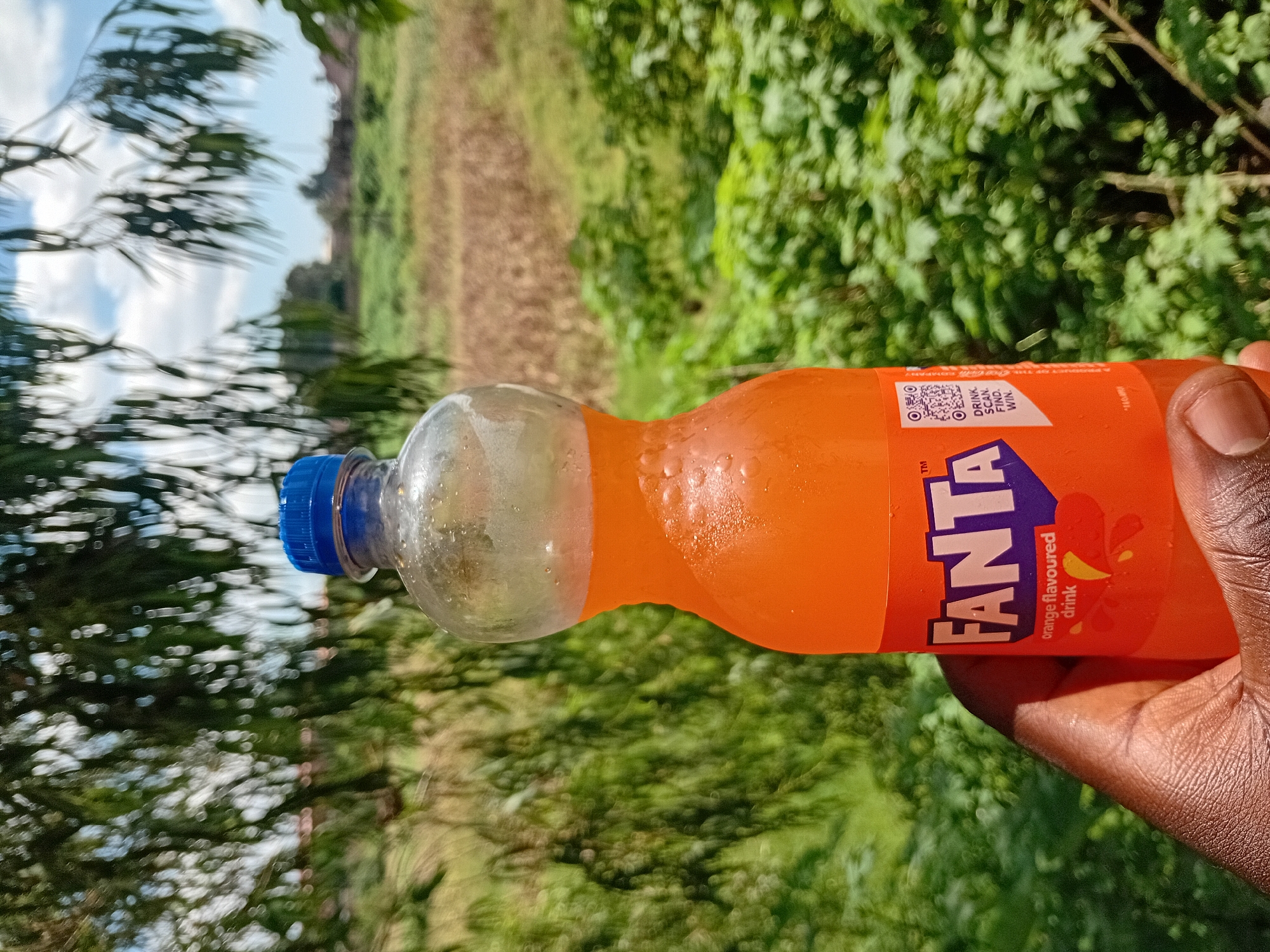 An outdoor, close-up, eye-level shot shows a dark-skinned person's left hand holding a clear plastic bottle of Fanta orange-flavored drink. The bottle is partially filled with orange liquid and has condensation on its surface. The label on the bottle features the Fanta logo in purple and white lettering, along with text that reads "orange flavoured drink." A QR code is also visible on the label. The background is blurred, with lush green foliage and a dry, grassy field under a partially cloudy sky. The setting appears to be Barakin Lerawi, Nigeria.