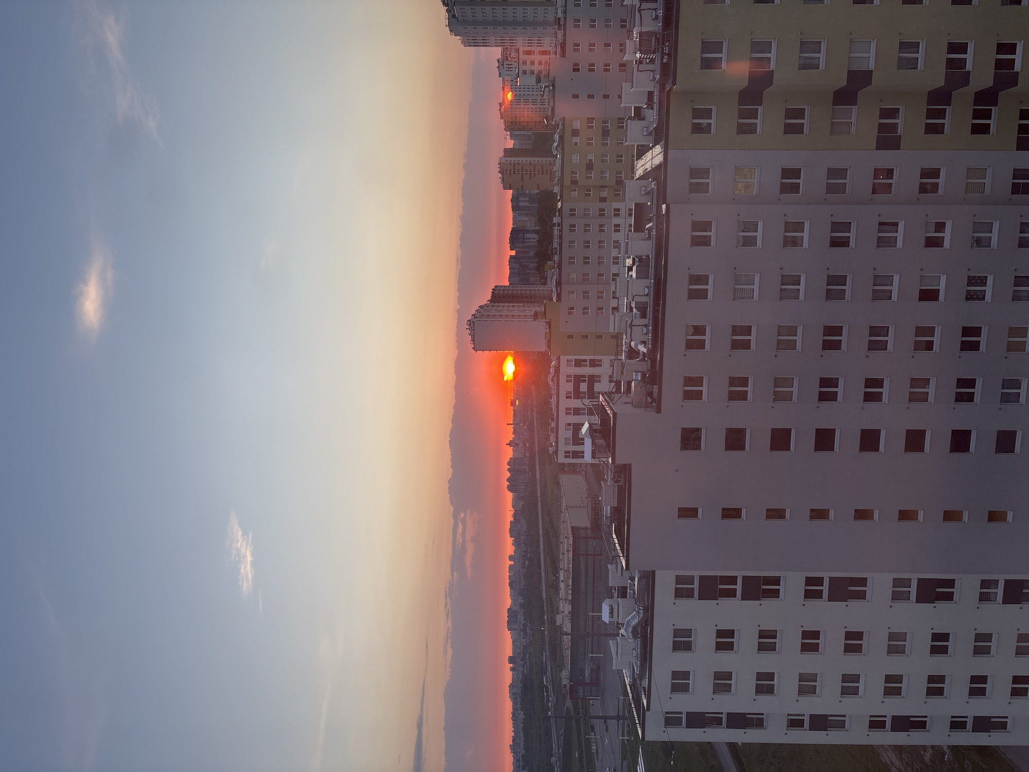 The image shows a cityscape at sunset, likely taken from a high vantage point. The setting sun is visible on the horizon, obscured by buildings and atmosphere, creating a bright orange glow in the sky. A tall residential building with multiple windows dominates the right side of the frame. Other buildings, including high-rises, are visible in the distance. The landscape below features trees, roads, and possibly industrial or commercial structures.
While the exact location within Vsevolozhsk cannot be determined without more specific landmarks or details, the scene appears to be from a residential area with a mix of high-rise and lower-level buildings.