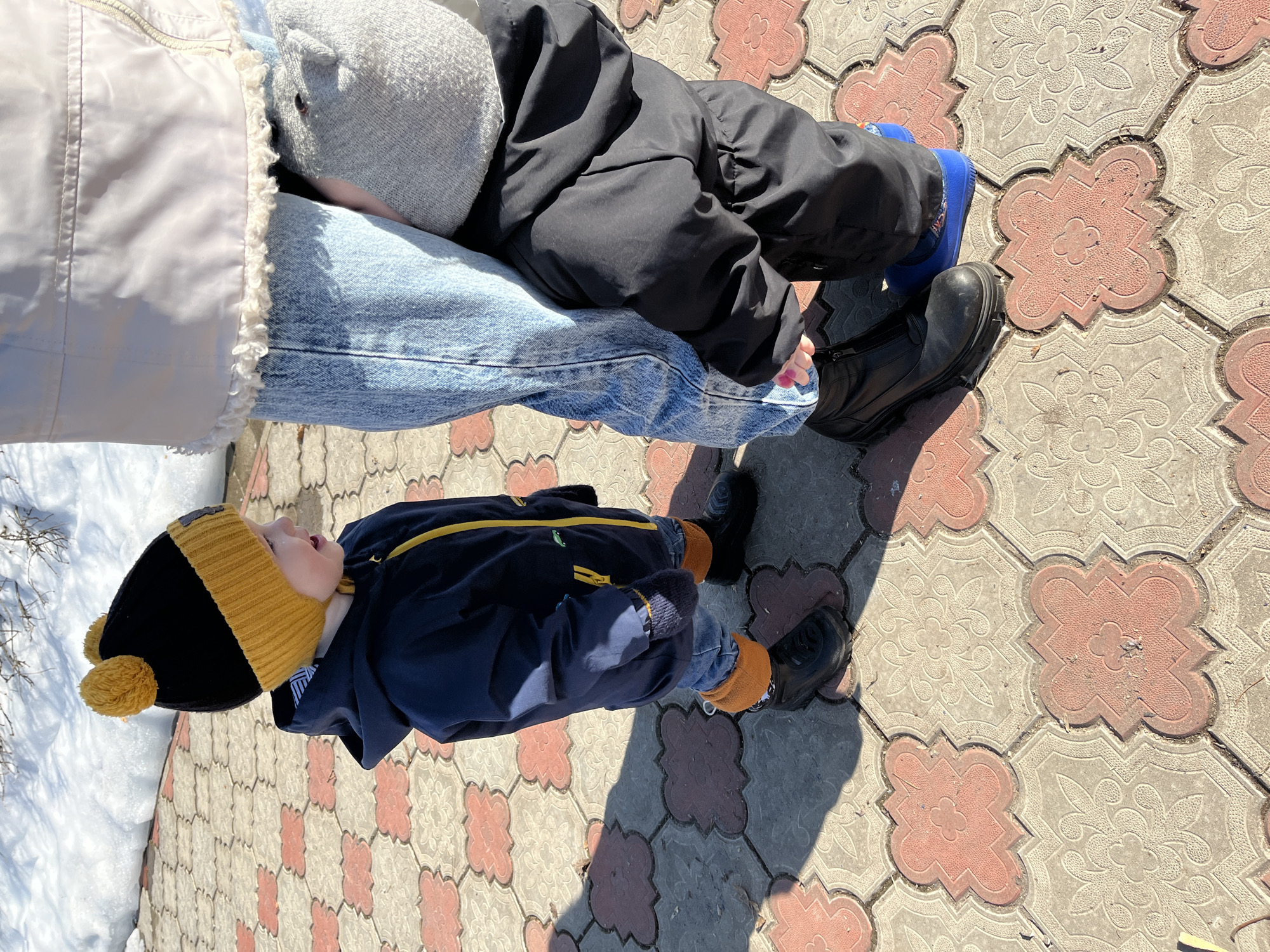 The image shows a person holding a child outdoors on a paved surface in the city of Chaikovsky, Russia. The adult is wearing jeans, a light-colored vest with a fleece lining, a gray hat, and a black winter coat. They are sitting with another child on their lap who is wearing a black coat, blue socks, and black boots. The child being held is dressed in a navy blue jacket with yellow trim, a black and yellow knit hat, and similar dark shoes. The paving stones are patterned in red and grey. Some snow is visible in the background.