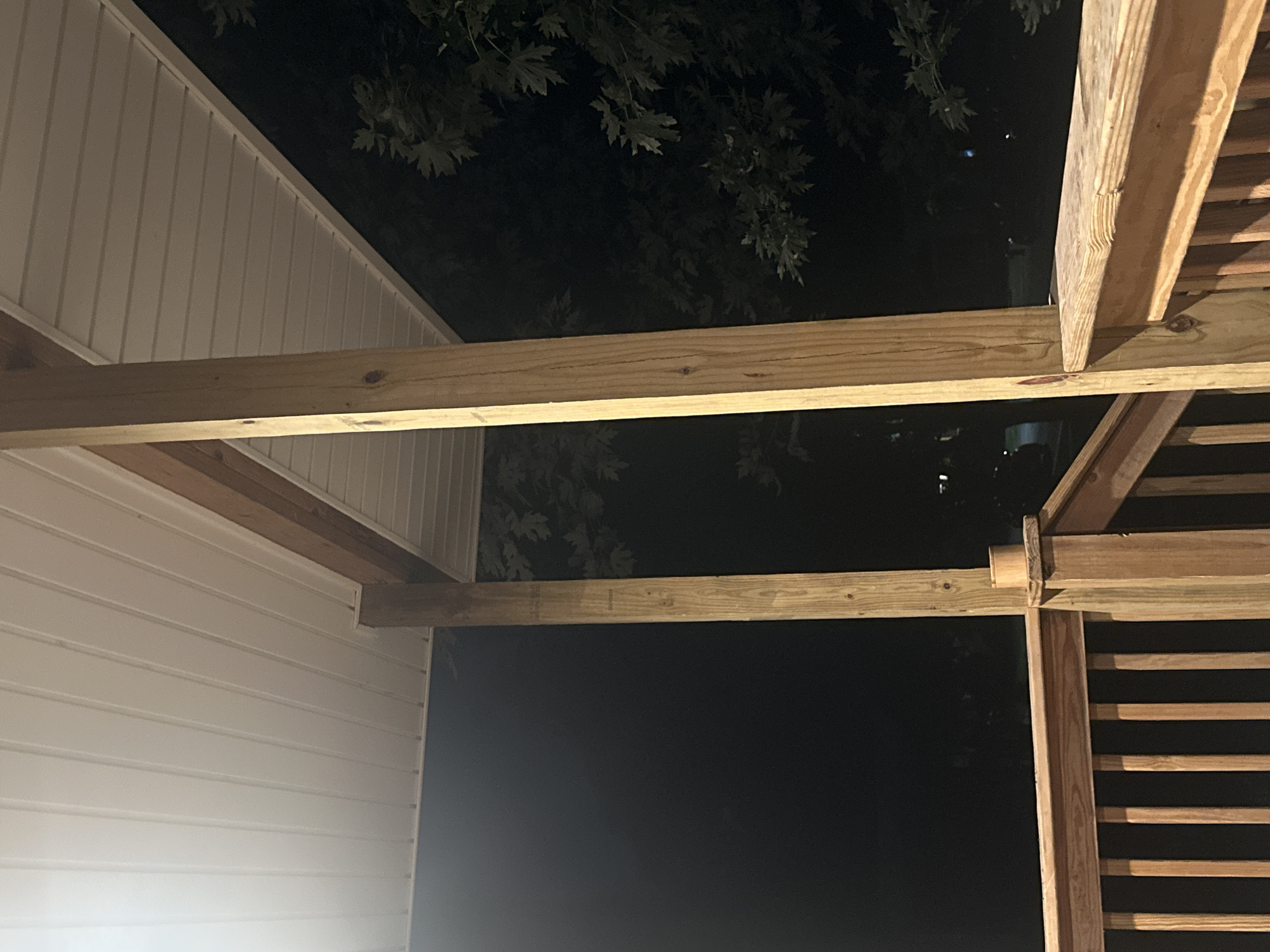 The image shows the underside of a wooden porch roof at night. The porch is supported by two vertical wooden posts and a horizontal wooden beam. The ceiling of the porch is white and paneled. In the background, there are dark trees with leaves and a dark sky. The porch railing is made of wood with vertical slats. The location is identified as Sedalia, United States of America. There are no people or animals visible in the image.