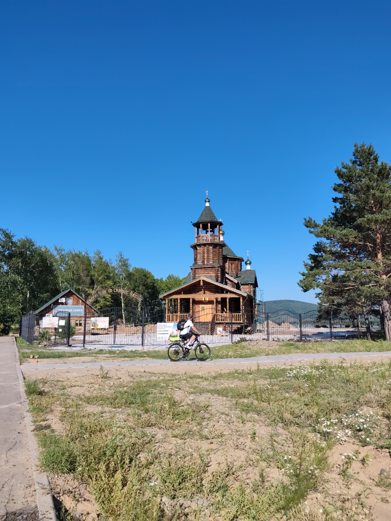 A wide, low-angle, outdoor shot shows a wooden Orthodox church with a bell tower and a domed roof, set against a clear blue sky. In the foreground, a person on a bicycle with a backpack rides on a paved path. The church is surrounded by a black metal fence, and to the left of the church, there is a smaller wooden building with signage. Behind the church and fence, a dense forest of deciduous trees and a tall pine tree are visible. Rolling, tree-covered hills can be seen in the distance. The ground in the immediate foreground is a mix of dirt and sparse vegetation, with some wildflowers scattered throughout. The scene appears to be a religious site or park, likely in a rural or semi-rural area.