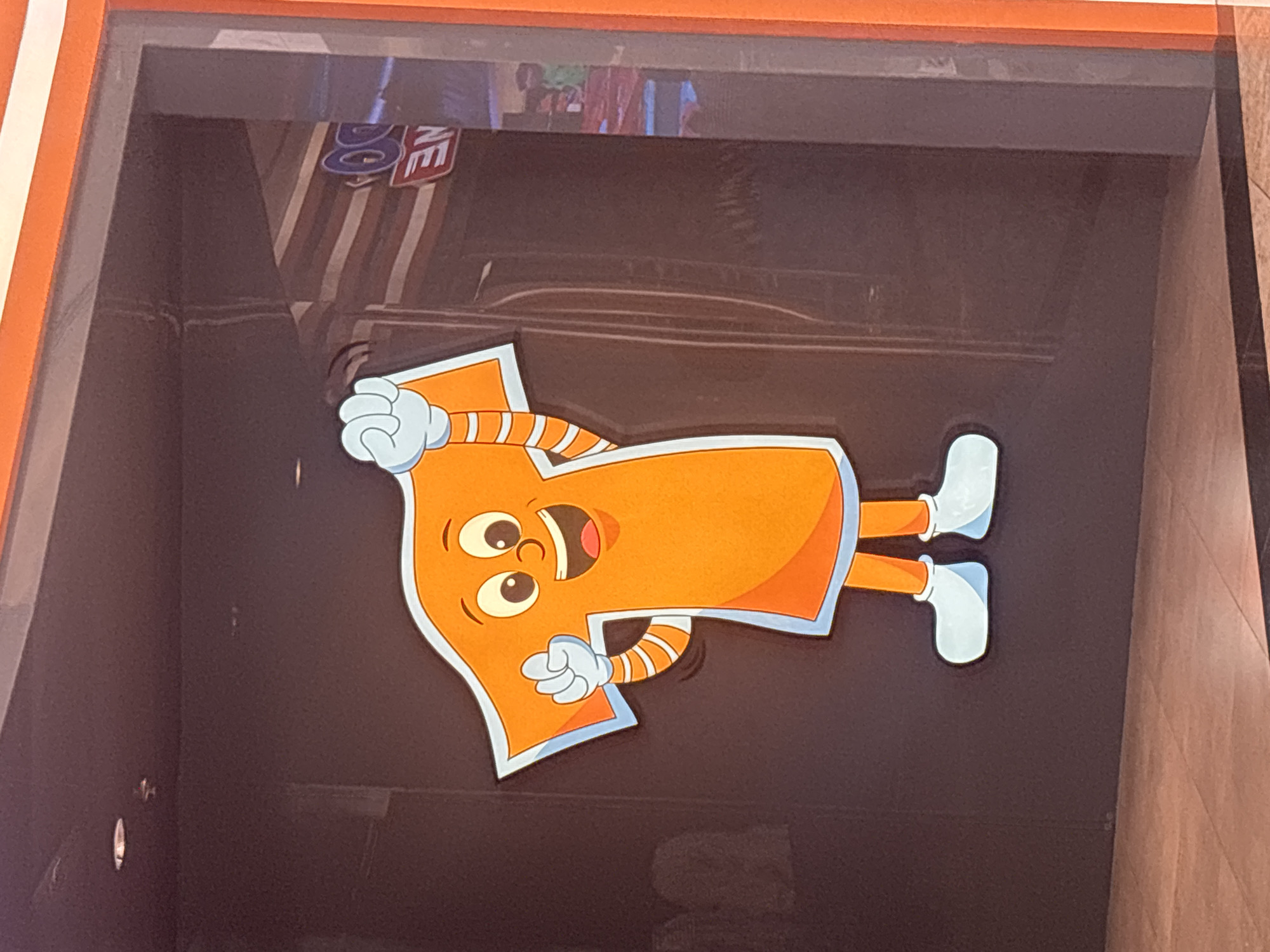 An orange cartoon character, shaped like the letter "T", is displayed on a dark, reflective surface. The character has large eyes, a wide smile, and is wearing white gloves and shoes. It is positioned in the foreground, with the background showing a blurred reflection of a store interior, including what appears to be signage with the letters "NE" and "GO". The character is the primary subject, with its animated pose suggesting a welcoming or playful gesture. The overall scene appears to be an indoor commercial space, possibly a shop or restaurant, given the reflective surfaces and interior details in the background. The setting is not definitively identifiable as Dubai, United Arab Emirates, as there are no specific landmarks or contextual clues in the image to confirm this location.