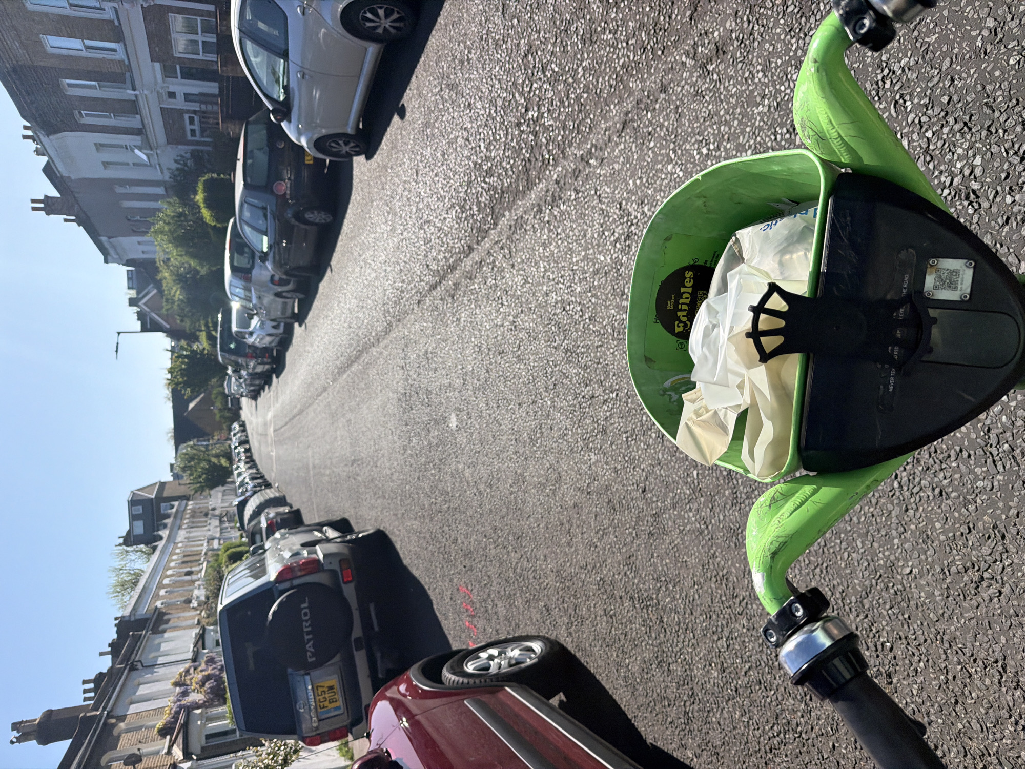 The image shows a street scene in London, United Kingdom, taken from the perspective of someone on a Lime scooter. The street is lined with parked cars on both sides, stretching into the distance. Buildings are visible behind the parked vehicles. The scooter's front basket is in the foreground, containing crumpled white material. The road surface is asphalt. The sky is blue. The license plate 'FG57 BUW' is visible on the silver Patrol car, parked on the left side of the street.