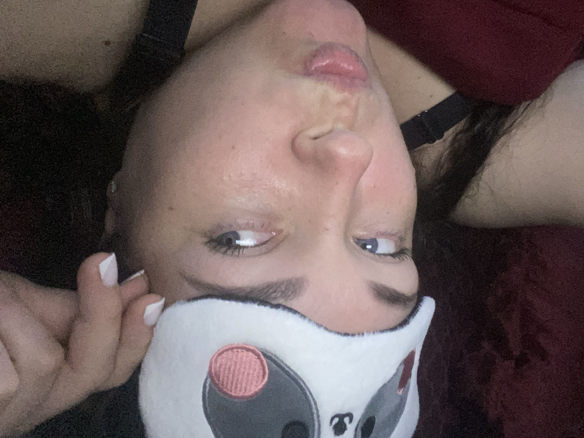 A woman is looking into the camera with a panda-themed sleep mask partially covering her face. She has light skin, blue eyes, and dark hair. Her lips are puckered slightly. Her hand is visible near her face, with white-tipped nails. She is wearing what appears to be a black strap, possibly from a bra or camisole, and a dark red top or garment. The image has been rotated, so she appears to be upside down. The location in the city of Sochi, Russia cannot be determined from the content of the image.