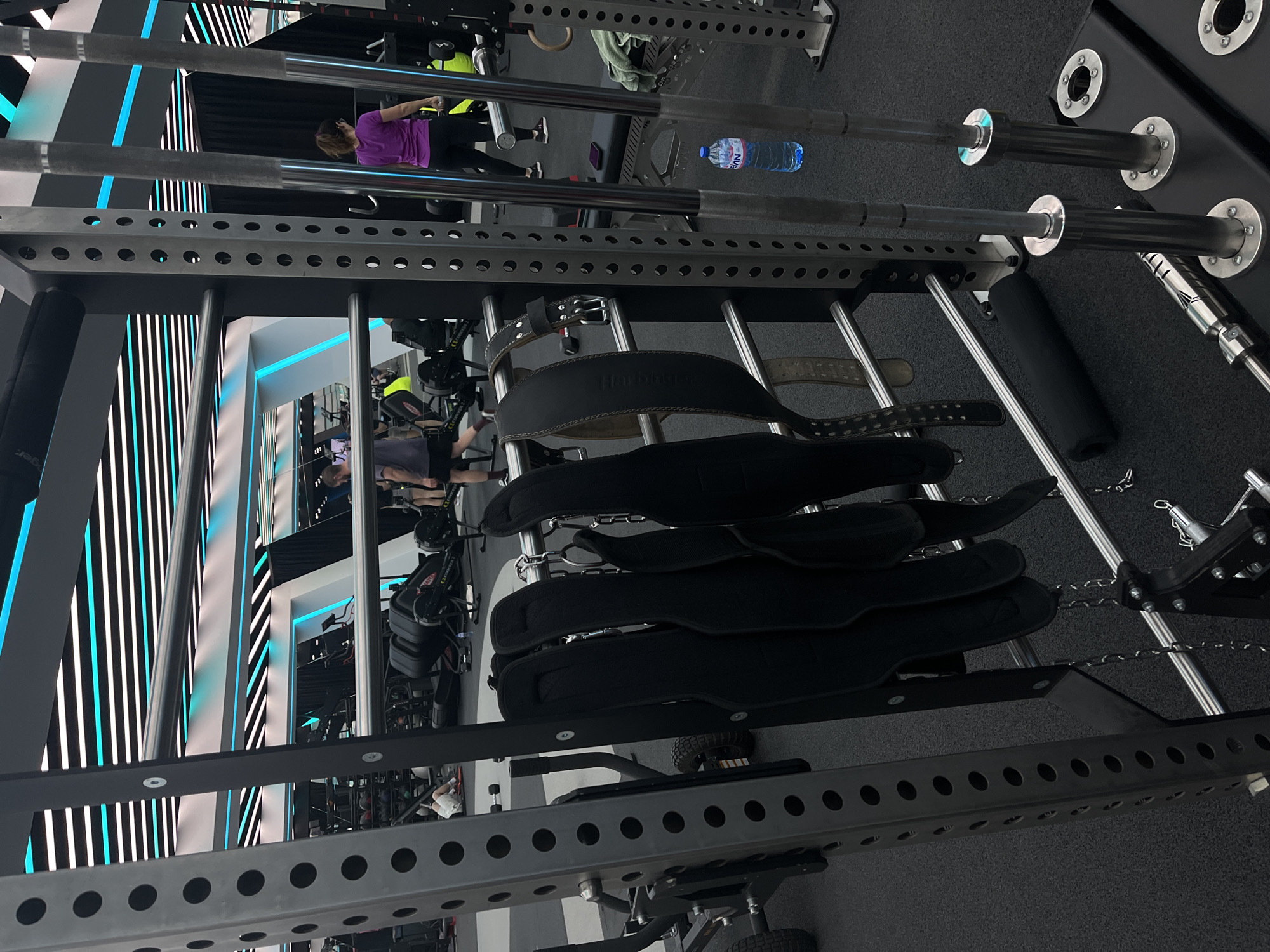 The image depicts an indoor gym environment. It shows a weight rack with several weightlifting belts hanging from it. In the background, people are exercising on various gym equipment. A water bottle is visible on a nearby weight bar. The gym has a modern design with black and white striped walls and a gray floor.