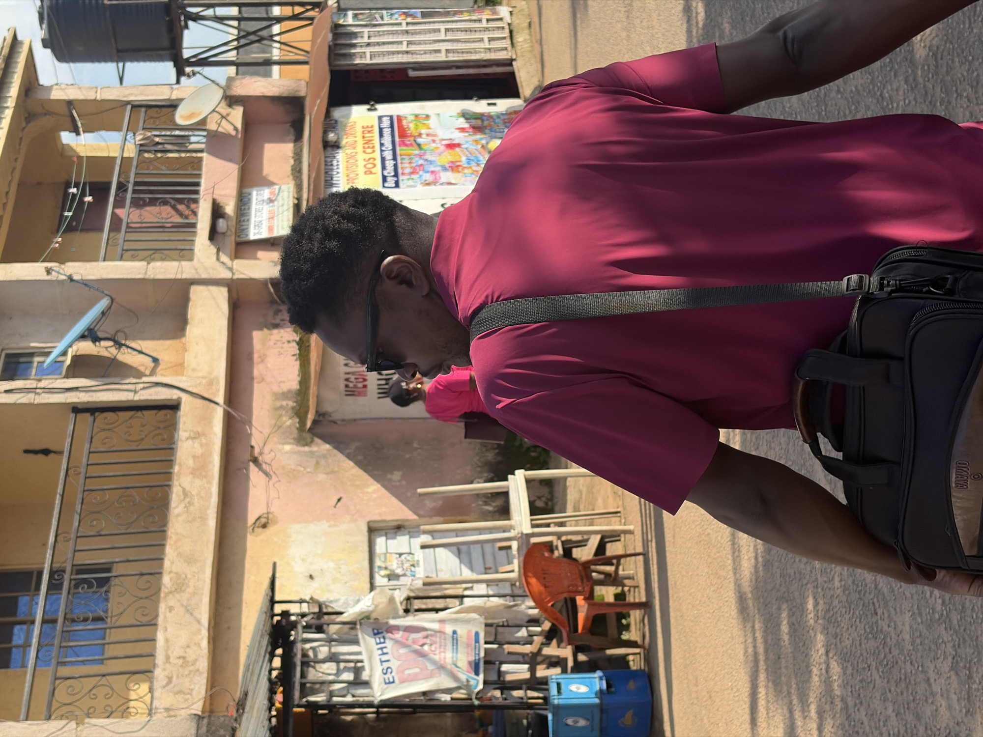 A man wearing sunglasses and a magenta shirt walks away from the camera, carrying a black satchel over his shoulder. He is walking on a dirt path in front of a row of buildings that appear to be shops. One building has a sign that reads "MEGA WHOLESALE PROVISIONS AND DRINKS POS CENTRE". There are chairs and stacked blue crates to the left of the man. The scene appears to be in Jos, Nigeria.