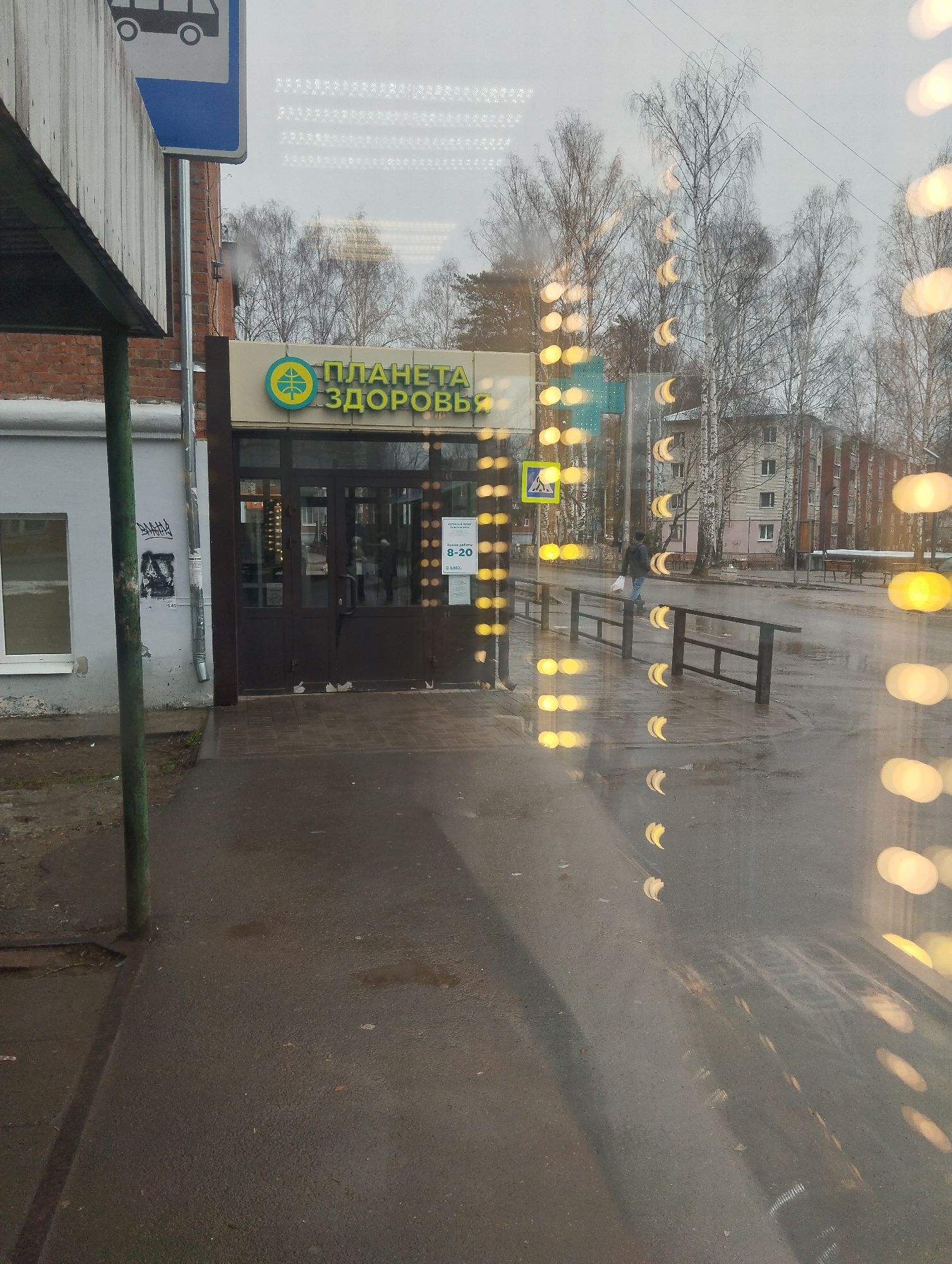 The image shows the exterior of a "Planet Health" pharmacy ("Planeta Zdorov'ya") in Votkinsk, Russia. The pharmacy has a brown entrance with glass doors. Above the entrance, a sign displays the pharmacy's name in Russian. To the left, a bus stop sign is visible. The foreground includes a wet sidewalk. Through the pharmacy's glass, a street scene is reflected, including trees, a pedestrian crossing the street, and an apartment building in the background. Bokeh lights appear throughout the image, possibly due to the camera's focus.