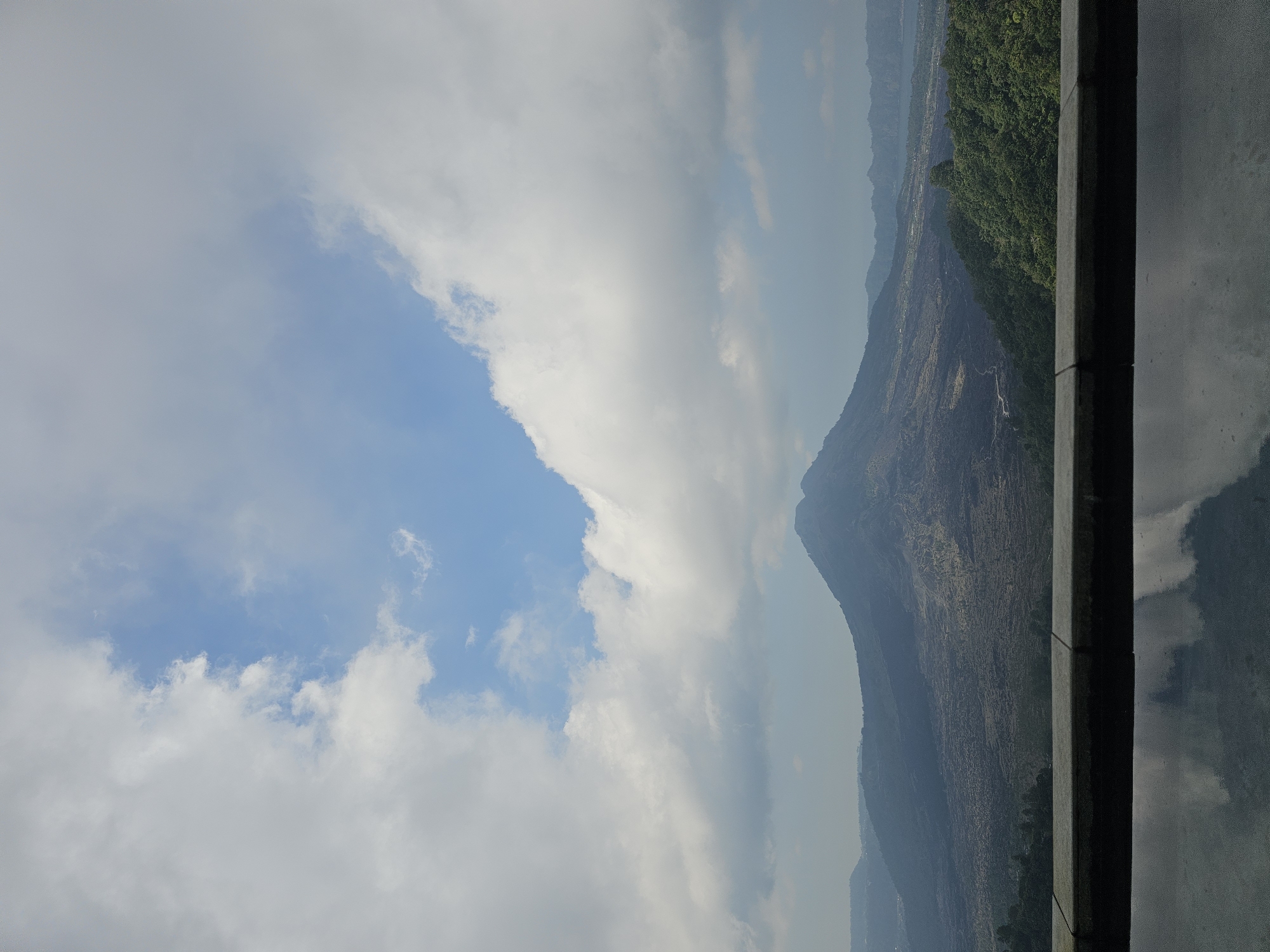 The image displays a panoramic view of Mount Batur, a volcano located in Kintamani, Indonesia. The foreground features a reflective surface, likely a pool of water or glass, mirroring the sky and the distant landscape. A stone ledge or wall runs horizontally across the lower portion of the image, separating the foreground from the main scene.
The middle ground is dominated by the imposing presence of Mount Batur, its slopes covered in a dark, rugged terrain that appears to be solidified lava. Beyond the volcano, a vast expanse of land stretches out, characterized by varied textures and vegetation. Rolling hills and a body of water, likely Lake Batur, can be discerned in the far distance. The landscape is predominantly green and brown, with some areas of darker, volcanic rock.
The sky above is a blend of bright blue and white clouds. Large, fluffy cumulus clouds are scattered across the sky, with patches of clear blue sky visible between them. The lighting suggests it is daytime, possibly late morning or early afternoon, with the sun casting a diffused light on the scene. There are no people or visible animals in the image. The overall impression is one of natural grandeur and tranquility.