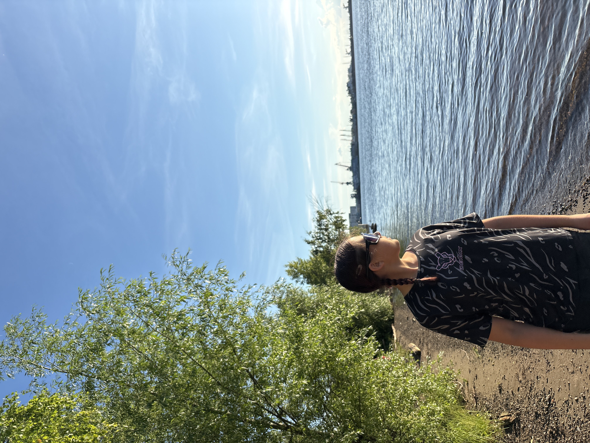 In this sideways, medium shot, a young person with braided brown hair and sunglasses stands by the water on a sunny day. They are wearing a black t-shirt with a grey animalistic pattern and the word "DEMIX" in purple. The person is looking off to the side, towards the water and distant cranes. The scene appears to be a waterfront area, possibly a port or industrial zone, with calm water reflecting the bright blue sky and scattered clouds. Lush green trees are visible in the foreground and on the bank. The lighting suggests it is daytime, likely late morning or early afternoon, given the strong sunlight and clear skies. The overall mood is relaxed and contemplative. The setting is suggestive of Chaikovsky, Russia, given the provided location context, possibly a riverbank or canal.