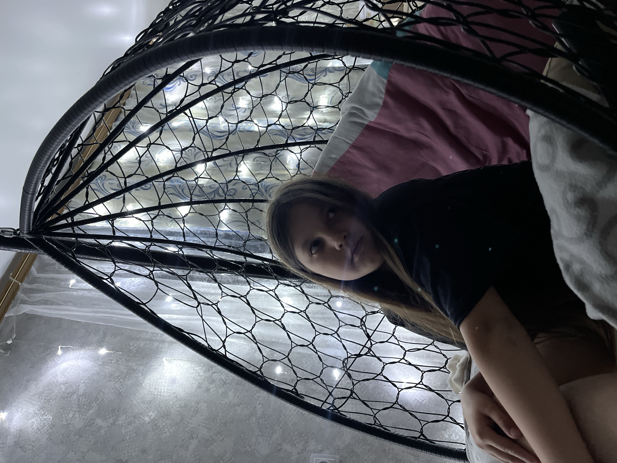 A girl lies in a hanging woven chair in an indoor setting, seemingly in a residential area in Novopokrovskaya, Russia. The chair, made of dark material, is adorned with string lights that shine through the woven structure. The girl is positioned sideways, looking up towards the camera with a neutral expression. She is wearing a dark-colored shirt. The background includes elements of the room's interior, such as a white ceiling, a window with white curtains, and wall decorations with lights. A pillow and a blanket with pink and white colors are seen in the background.