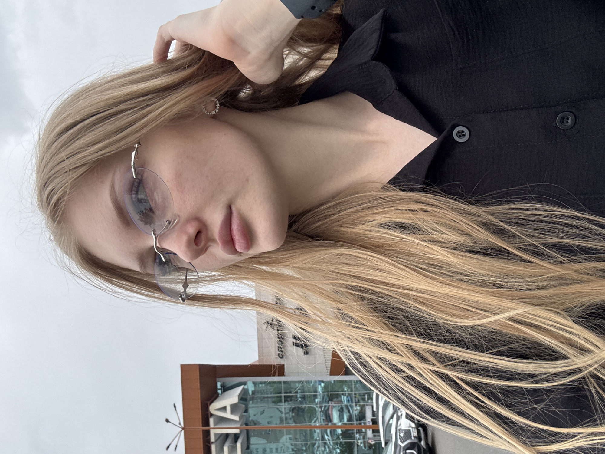 An outdoor, medium shot photograph shows a young blonde woman with fair skin and light freckles, wearing a black collared shirt and rimless oval-shaped glasses. She is looking to her left, her right hand raised to touch her hair. A circular earring with clear stones is visible on her right earlobe. In the background, a modern building with the Cyrillic letters "ETA" prominently displayed in white on its facade is visible. To the right of the "ETA" building, another building has Cyrillic lettering that appears to read "СПОРТ", suggesting it is a sporting goods store or complex. The buildings are part of a commercial area in the city of Perm, Russia, with cars parked in a lot in front of them. The sky is overcast.
