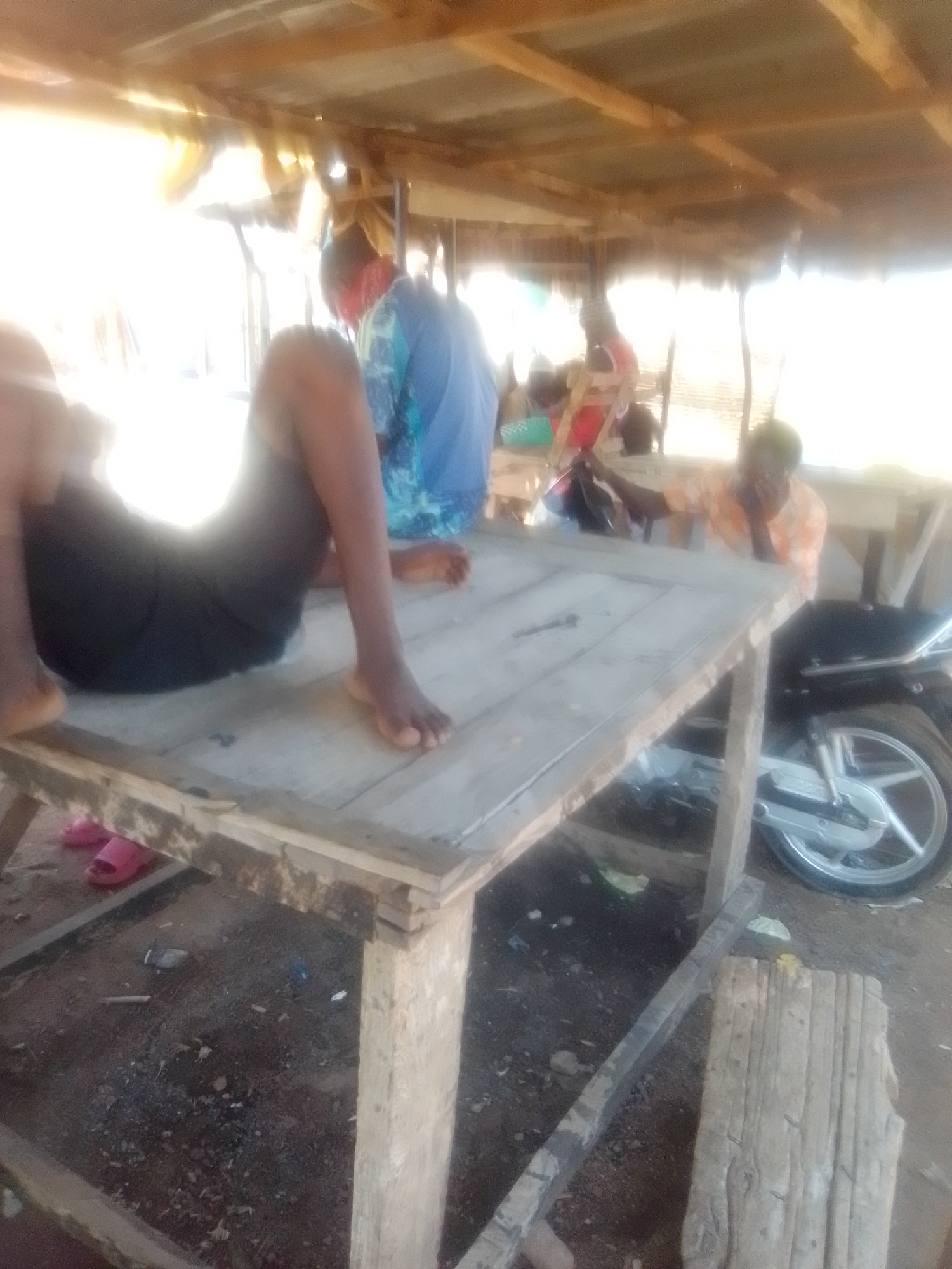 The image depicts an outdoor scene under a rustic, wooden shelter, possibly in Pantame, Nigeria. In the foreground, a person wearing black shorts and bare feet is partially visible, seated on a rough wooden table. Their legs are bent, with one foot resting on the table surface. Pink flip-flops are visible on the ground beneath the table.
Behind the table, another person is seen in profile, wearing a light-colored, patterned shirt and glasses. They are leaning on the table, with their right arm bent and resting on their head, appearing to be in a relaxed or contemplative pose. A motorcycle with a silver spoked wheel is parked to the right of this person.
Further in the background, other individuals are present, though less clearly defined due to the lighting and image quality. One person is wearing a blue and green patterned garment, and another is seen in an orange shirt. The environment is characterized by loose dirt, pebbles, and scattered debris on the ground. The shelter's roof is constructed of corrugated metal supported by wooden beams. The overall impression is of a casual, informal gathering in an open-air setting.