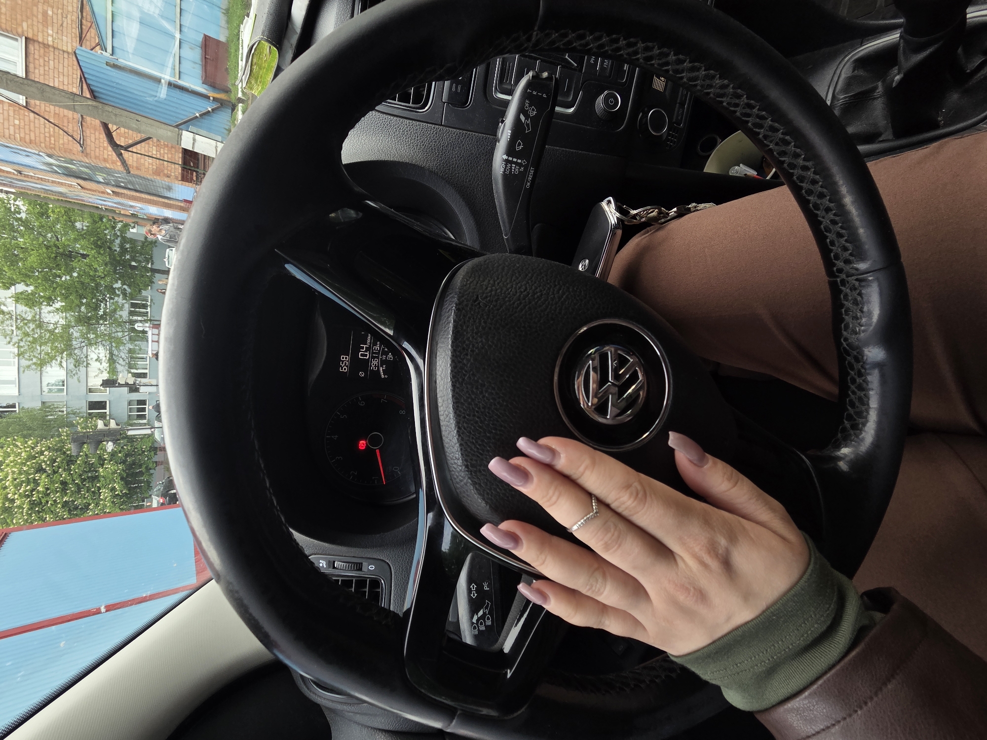 The image shows the interior of a car, focusing on the steering wheel and the hand of the driver. The car appears to be a Volkswagen, judging by the logo on the steering wheel. The dashboard and instrument panel are visible behind the steering wheel, displaying the time as 6:58 and some driving statistics.
A hand with long, mauve-colored nails is resting on the steering wheel. The person is wearing a dark green long-sleeved shirt, with the sleeve of a brown jacket visible. The keys are visible on the steering column next to the steering wheel,
Outside the car, through the windshield, a city scene is visible with buildings and trees. The exterior buildings appear to be made of blue siding and red brick.
The general location appears to be in a populated urban setting, possibly in the city of Unknown, Unknown.
