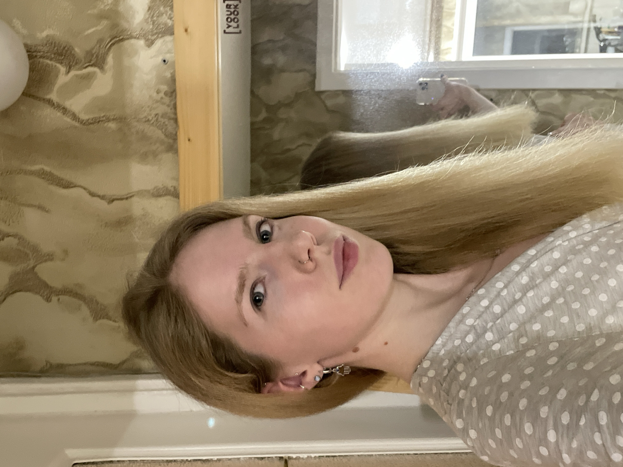 A Caucasian woman with blonde hair and blue eyes poses in front of a mirror. She has a nose ring and multiple earrings in her left ear. She is wearing a gray and white polka-dot top. The background includes a wall with textured wallpaper, a wooden frame, and a window. The reflection in the mirror shows a person holding a phone, presumably taking the picture. The location in Izhevsk, Russia cannot be determined from the image.