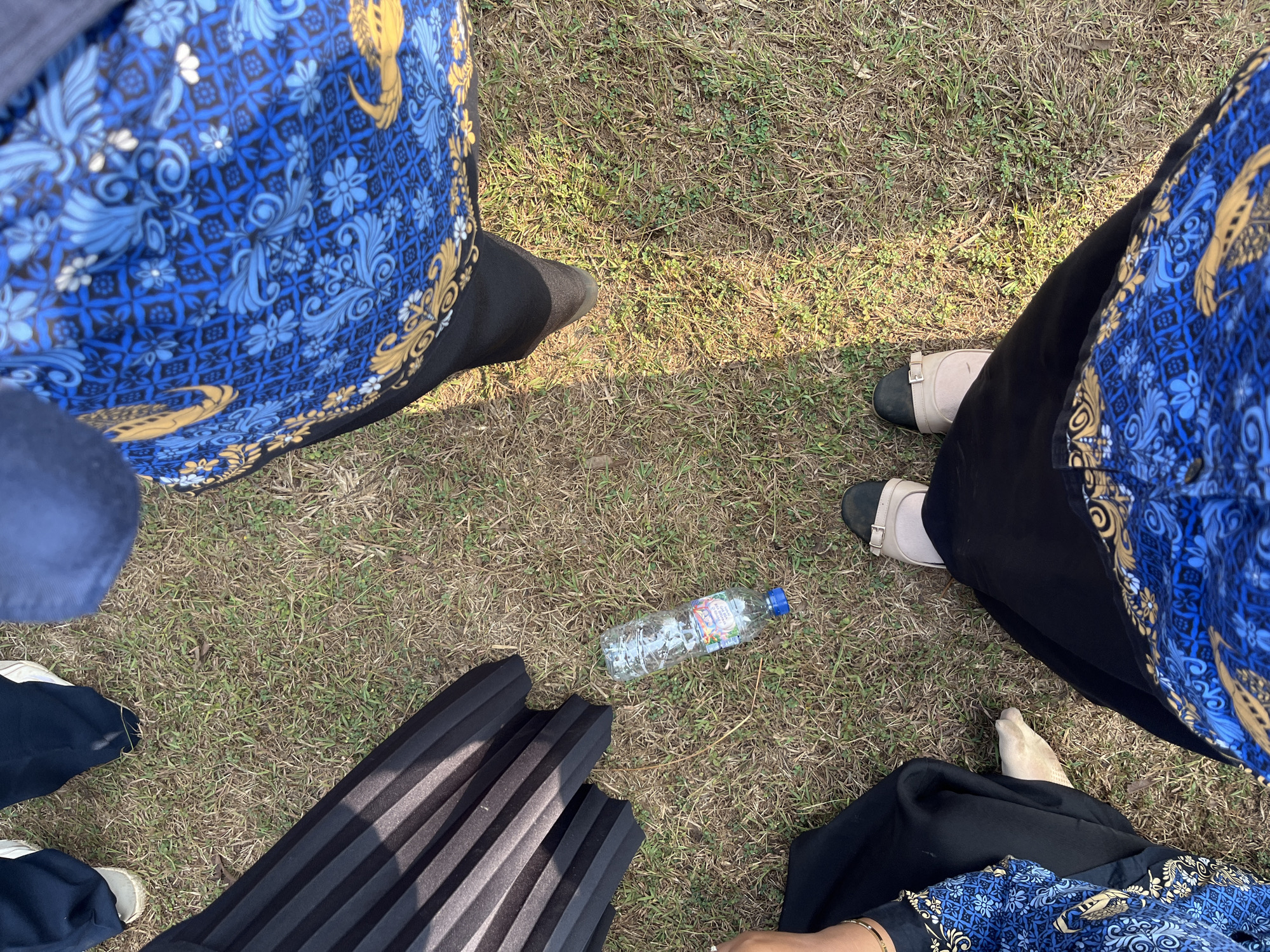 In Gembong, Indonesia, on a grassy outdoor area, several individuals are partially visible from a high-angle perspective. The ground is covered in dry grass, with some patches of green. A transparent plastic water bottle with a blue cap lies on the grass. One person is wearing black pants and light-colored shoes. Another person is wearing a blue and gold patterned garment, black pants, and light-colored shoes. A third person is wearing a pleated black skirt or garment. A fourth person is visible wearing a blue garment with a yellow and blue intricate pattern. The overall scene suggests a casual gathering or outdoor event.