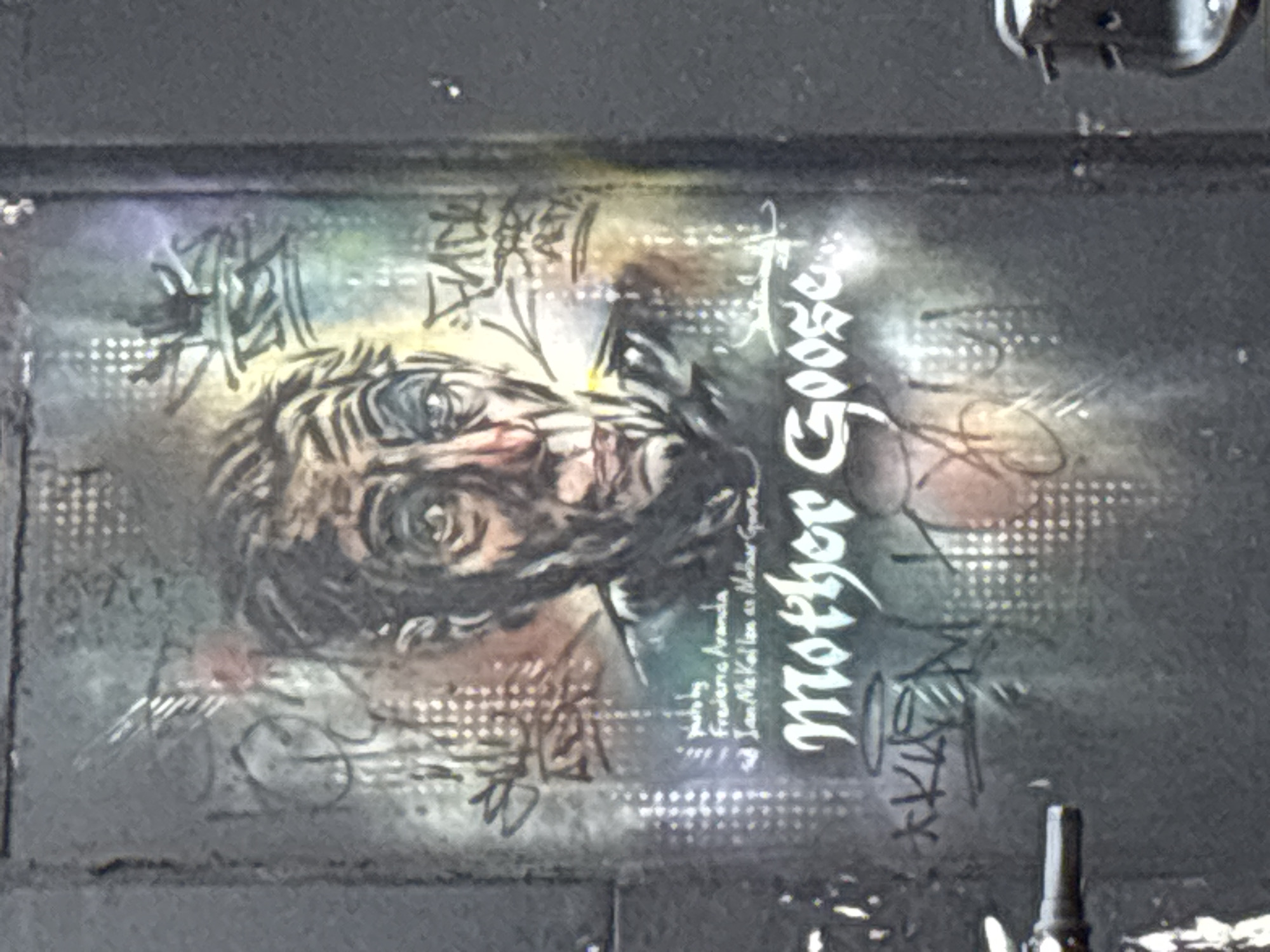 The image shows a graffiti-style artwork on a dark, possibly metallic surface. The artwork depicts a portrait of a person with heavily stylized features, surrounded by abstract shapes and lettering. The words "Mother Goose" are prominently displayed in a stylized, gothic font. Below the portrait, smaller text mentions "Frederic Aranda" and "Ian McKellan as Mother Goose". The environment appears urban, possibly an alleyway or industrial area, based on the dark, textured surface and presence of mechanical or architectural details at the top and bottom of the frame. The image was taken in the city of London, United Kingdom.